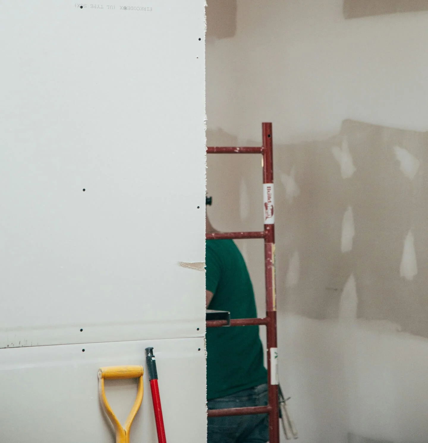Partially completed drywall installation on a construction site with visible seams and drywall patches, a worker in a green shirt working on the wall, and construction tools including a yellow dustpan and red-handled tool leaning against the wall.