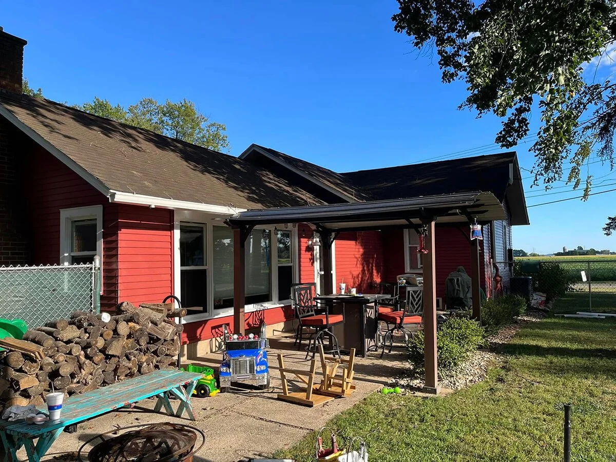 Backyard patio area of a red house with wood pile, children's toys, and outdoor furniture under a metal roof, with trees and open fields in the background.