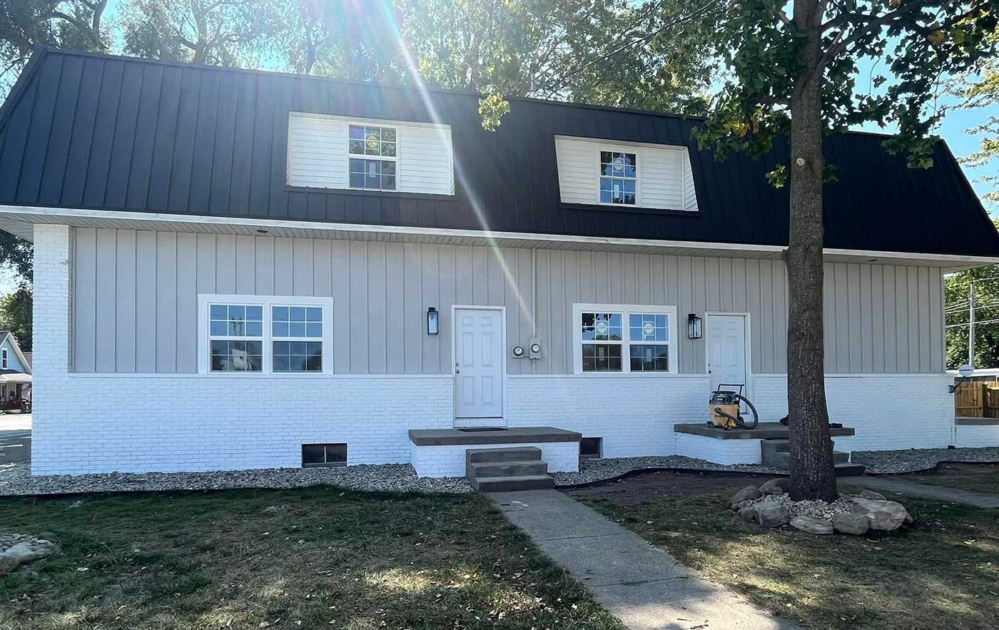 Newly constructed two-story house with white brick foundation, beige vertical siding, and a black metal roof. It has a front door with steps, two windows on the ground level, and two dormer windows on the upper level. There is a large tree in the front yard, and a sidewalk leading to the front door.