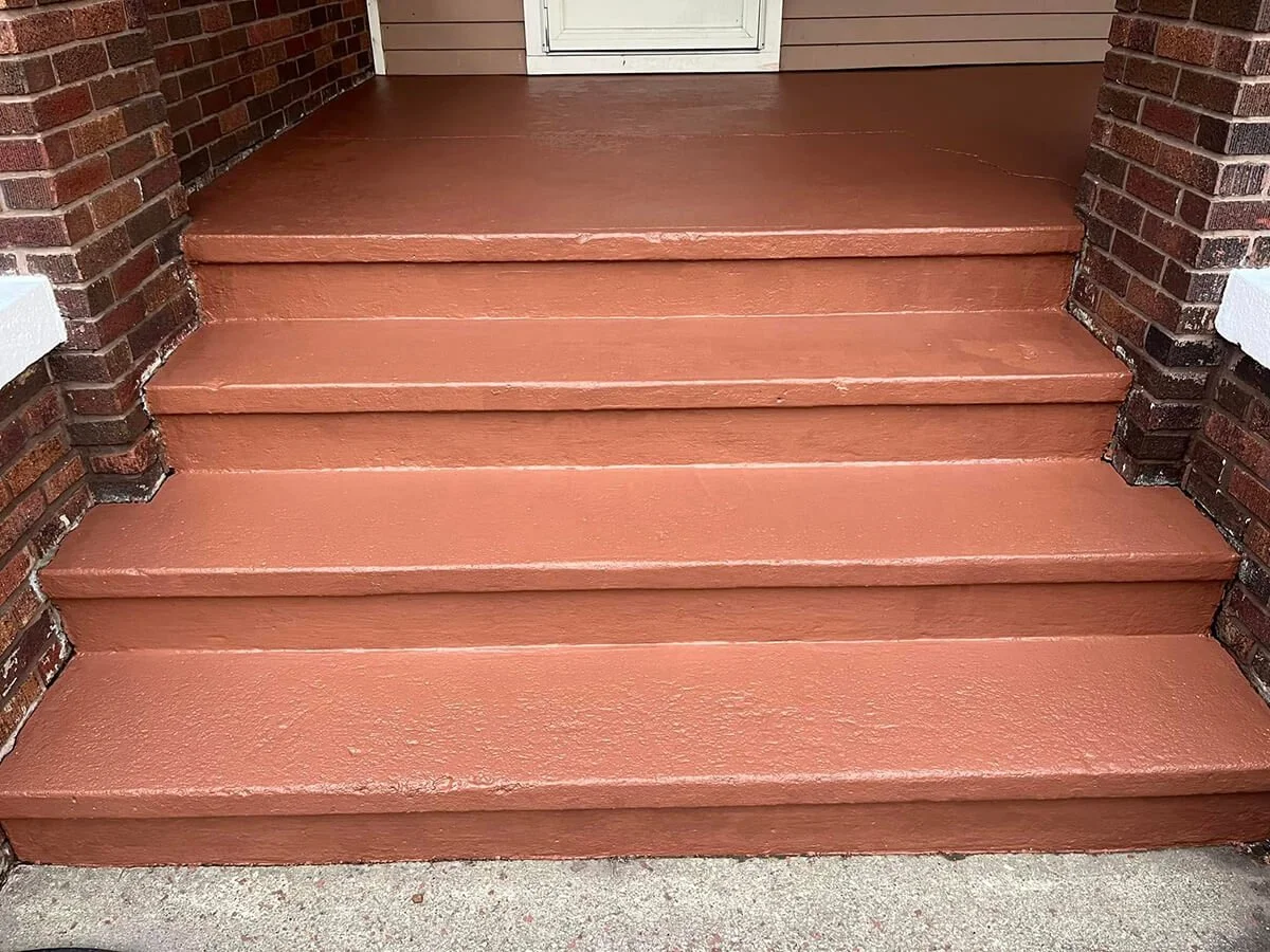 The image shows a set of four concrete steps painted in a reddish-brown color leading up to a white front door, with brick walls on either side.