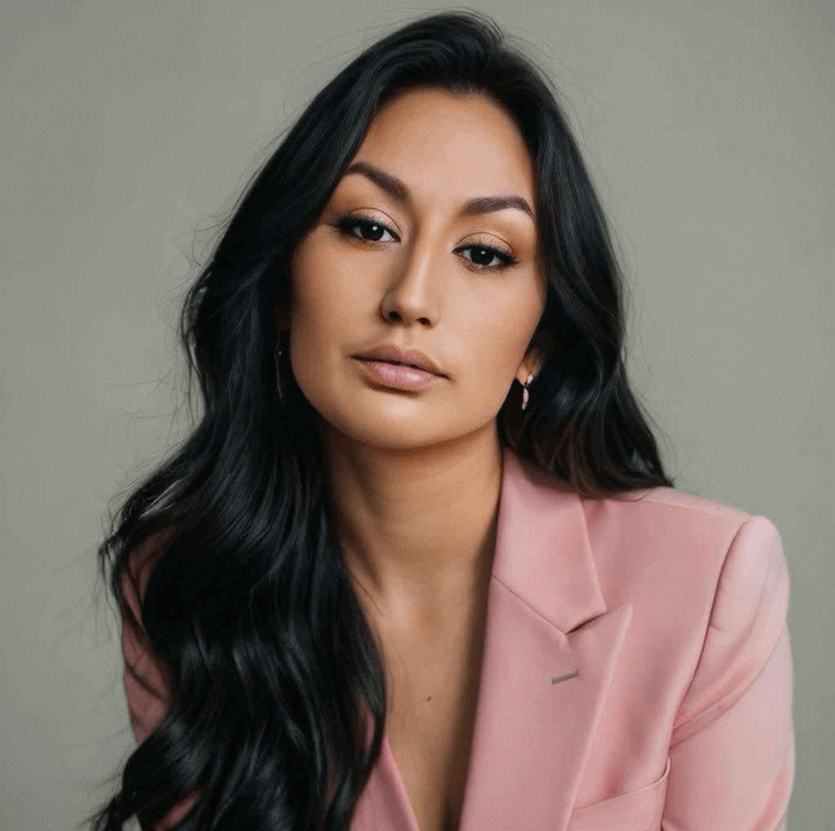 A woman with long, dark wavy hair, wearing a light pink blazer, looking at the camera with a neutral expression against a plain background.