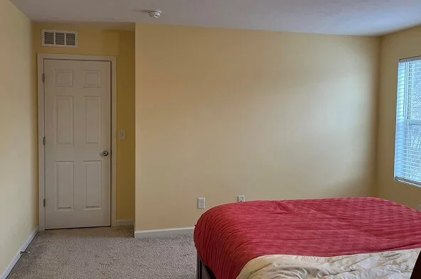 Empty room with yellow walls, a door, a window with blinds, and a bed with a red and beige comforter.