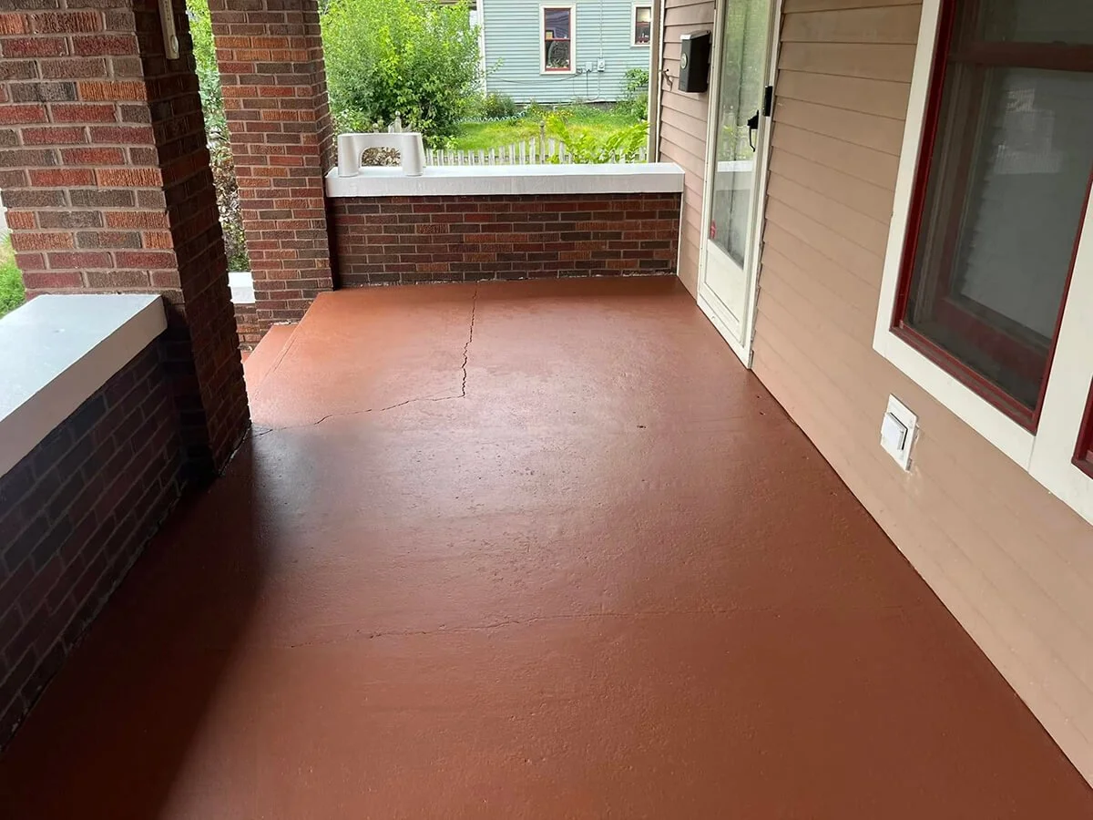Empty front porch with reddish-brown concrete flooring, brick walls, glass door with white frame, and view of green trees and neighboring houses in the background.
