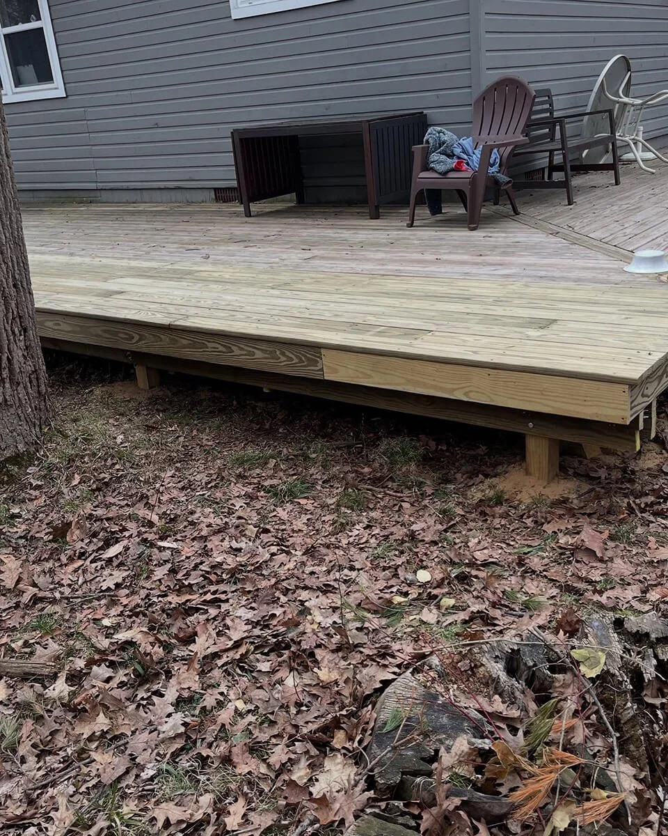 A newly built wooden deck attached to a house, with outdoor chairs and a table, overlooking a ground covered with fallen leaves and a tree trunk on the left.