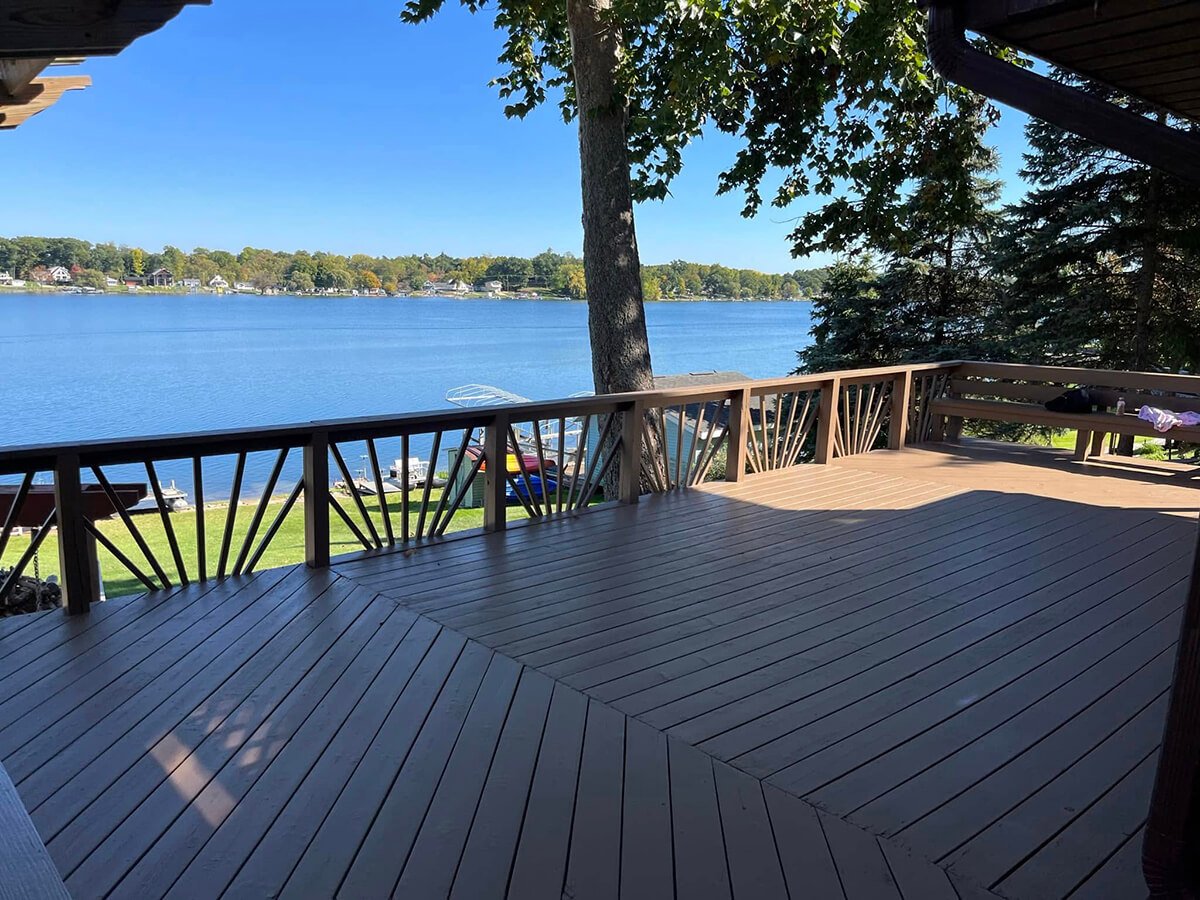 Wooden deck overlooking a lake with boats, trees, and houses in the distance on a sunny day.