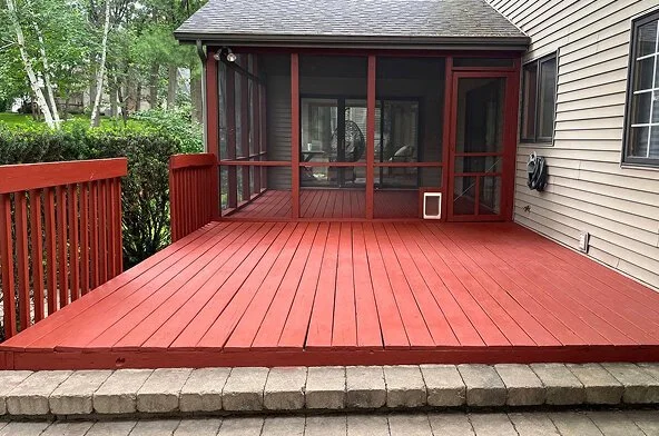 Red wooden deck attached to a house with a screened-in porch and patio furniture inside, surrounded by greenery.