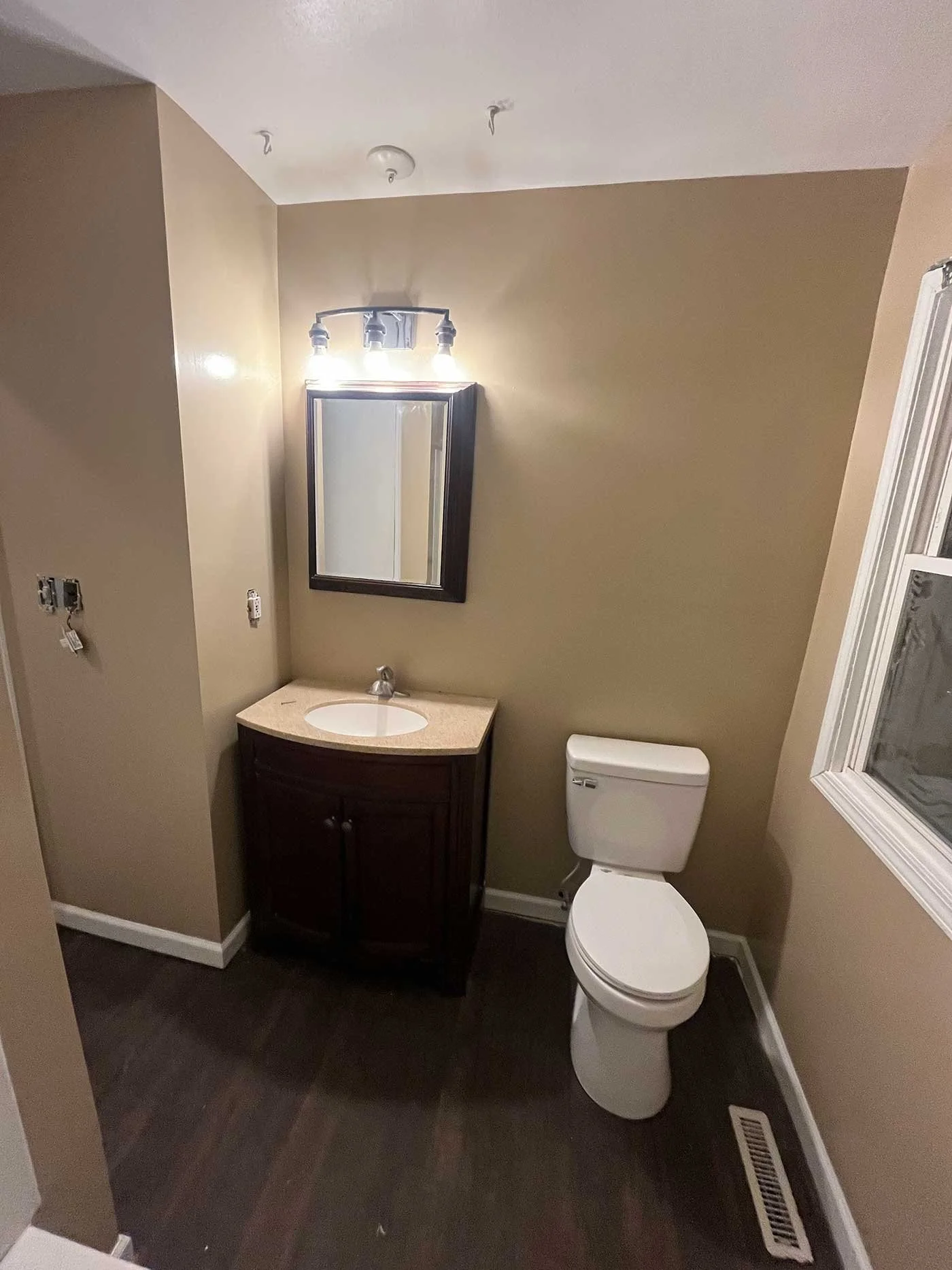 A small bathroom with a beige wall, a white toilet, a dark wood vanity with a beige countertop, a rectangular mirror, and a light fixture above the mirror. There is a window on the right side, and a vent on the floor near the wall.