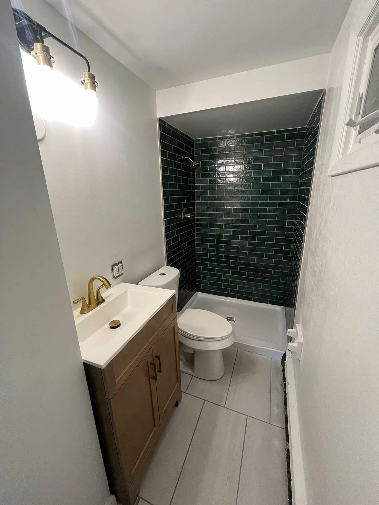 Small bathroom with a wooden vanity, a white sink with a gold faucet, a toilet, and a shower area with black tiled walls.