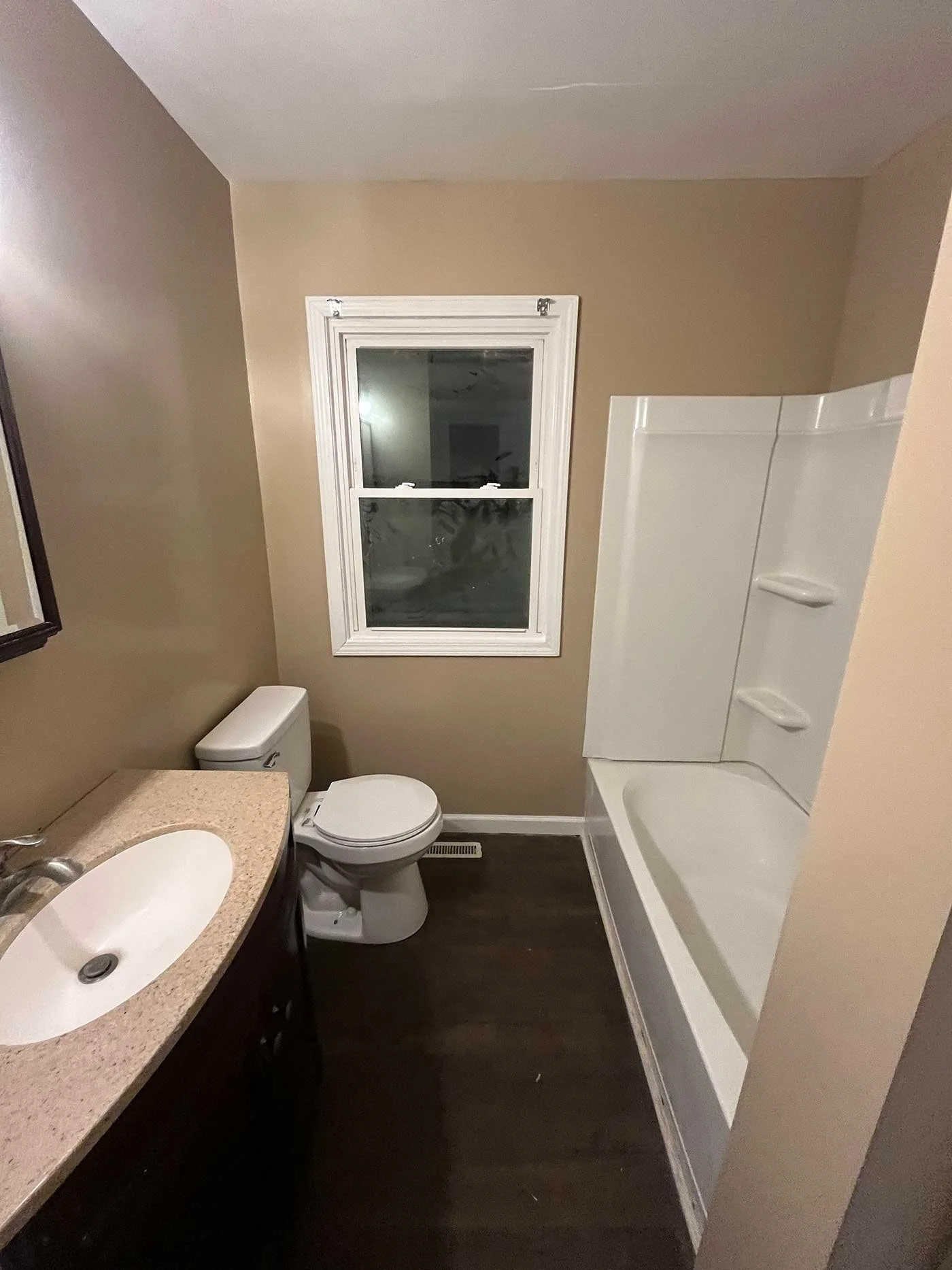 A small bathroom with beige walls, a single window, a white bathtub with a built-in shower, a toilet, and a dark-colored vanity with a beige countertop and an under-sink basin.