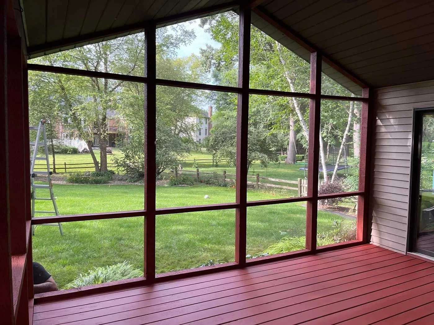 View from inside a screened porch looking out onto a green backyard with trees, a lawn, and neighboring houses in the distance.