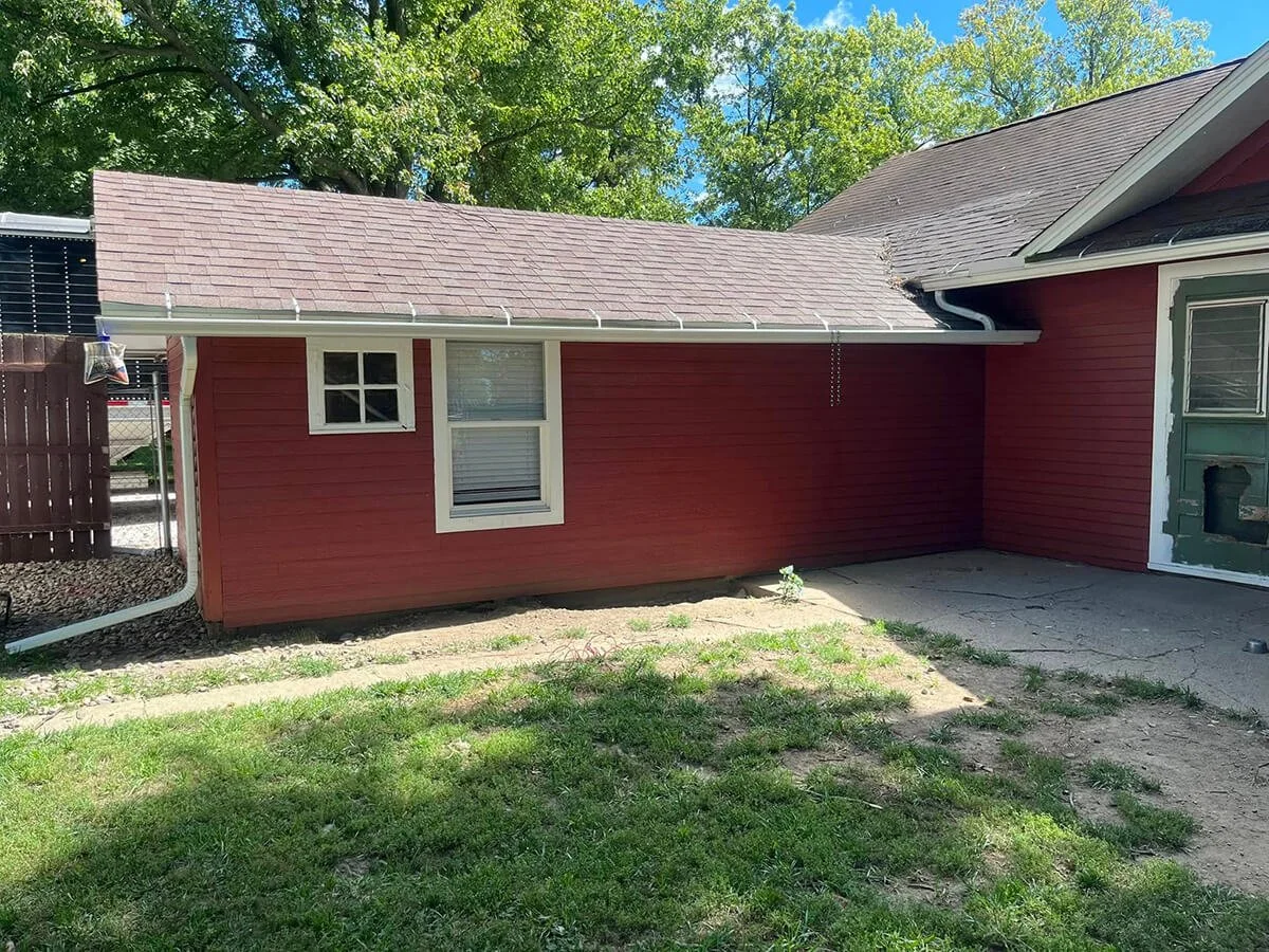 Backyard view of a red house with white trim, two small windows, a concrete patio, and a wood fence. The yard has patches of grass, and trees are visible in the background.