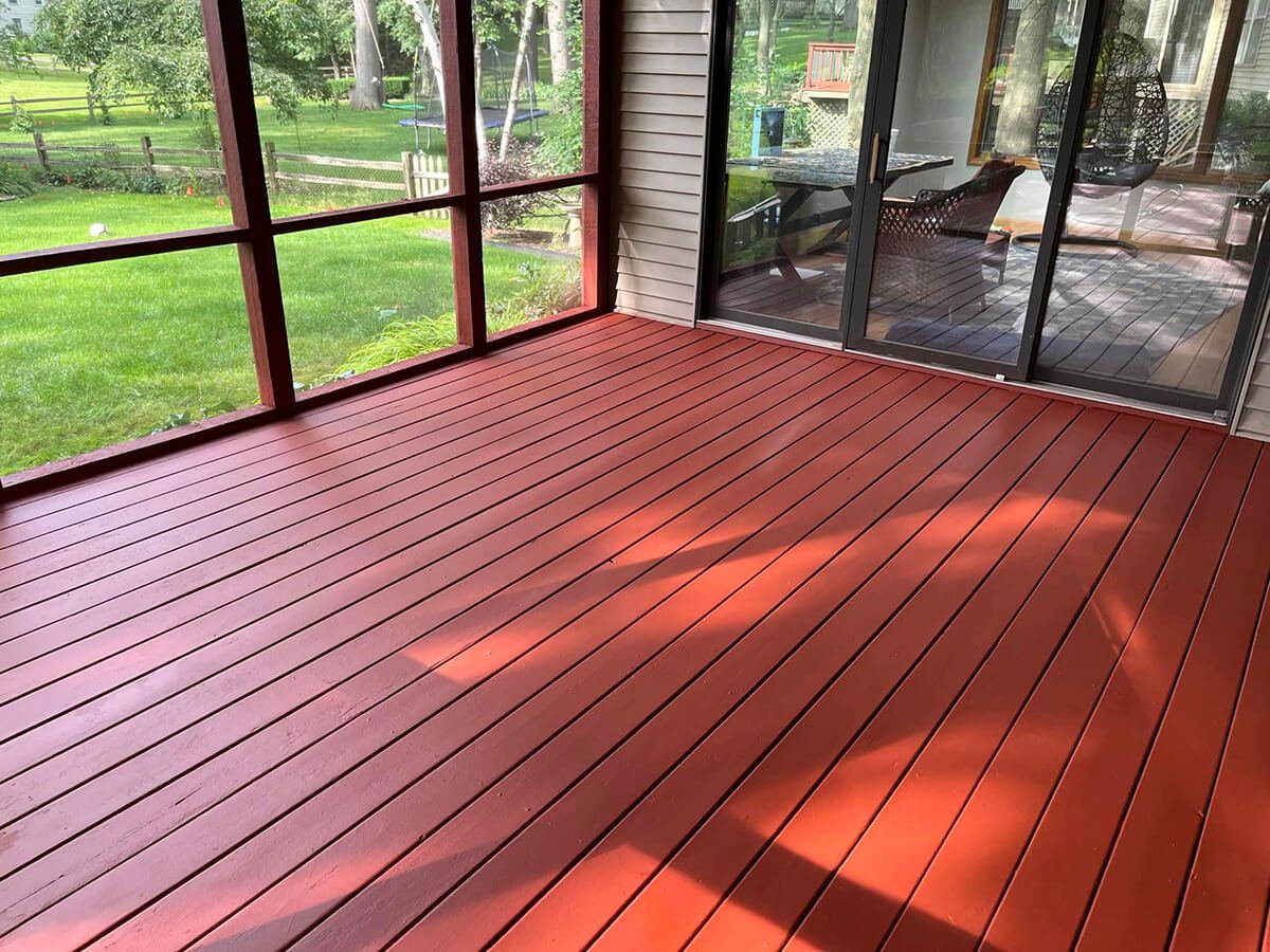 A screened porch with red wooden flooring and sliding glass doors leading to a backyard with green grass and trees.