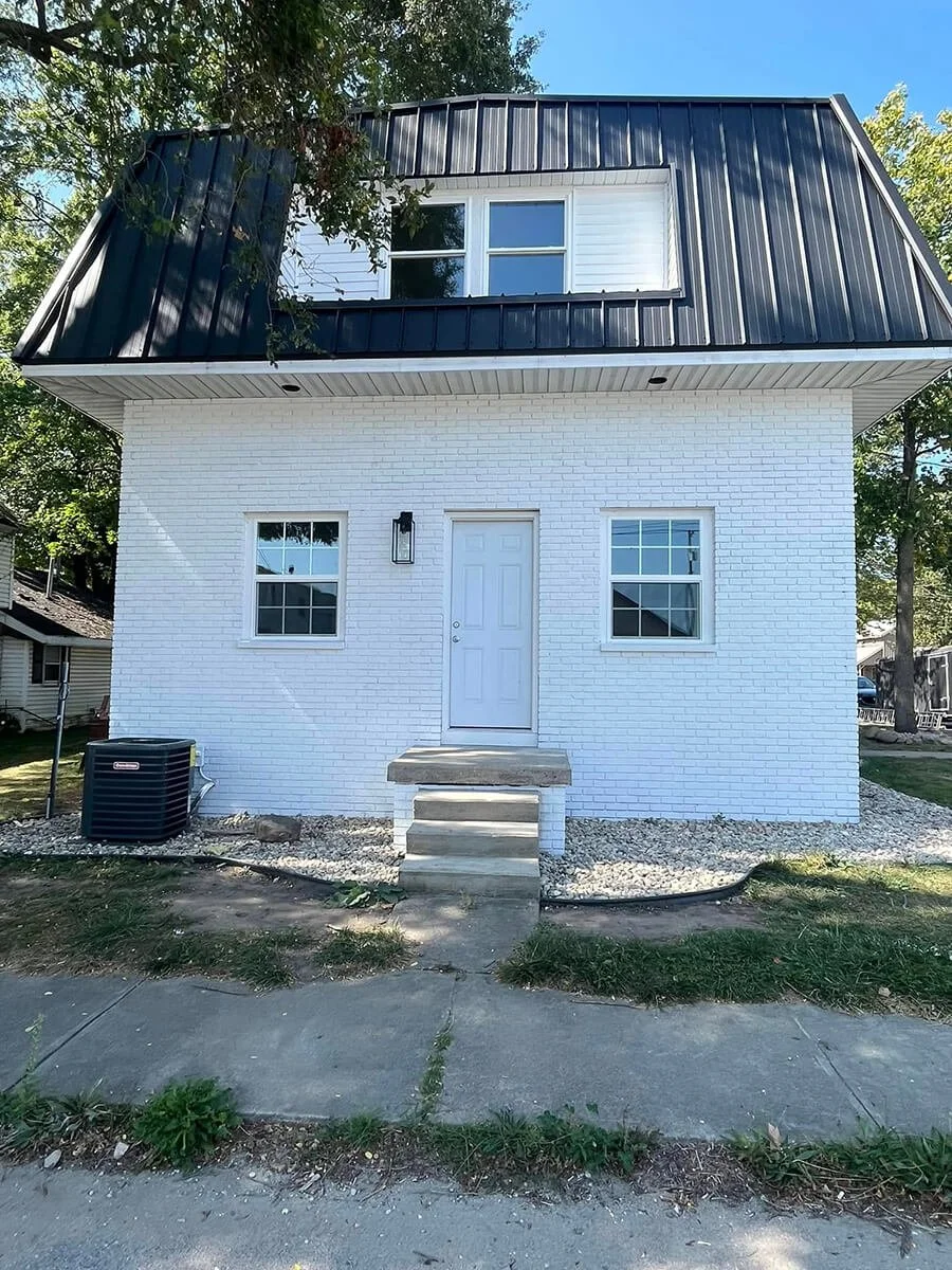 A two-story house with a white brick exterior on the lower level and white siding on the upper level, featuring a black metal roof, two windows on the lower level, a door with steps in the middle, and a horizontal window on the upper level. There is an air conditioning unit outside on the left and a sidewalk in front.
