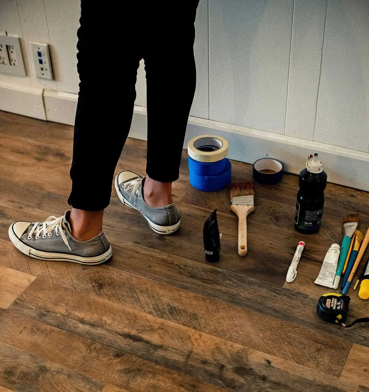 Person standing in front of a wall with various painting and tool supplies on the floor, including paint rollers, brushes, tape, and containers of paint.