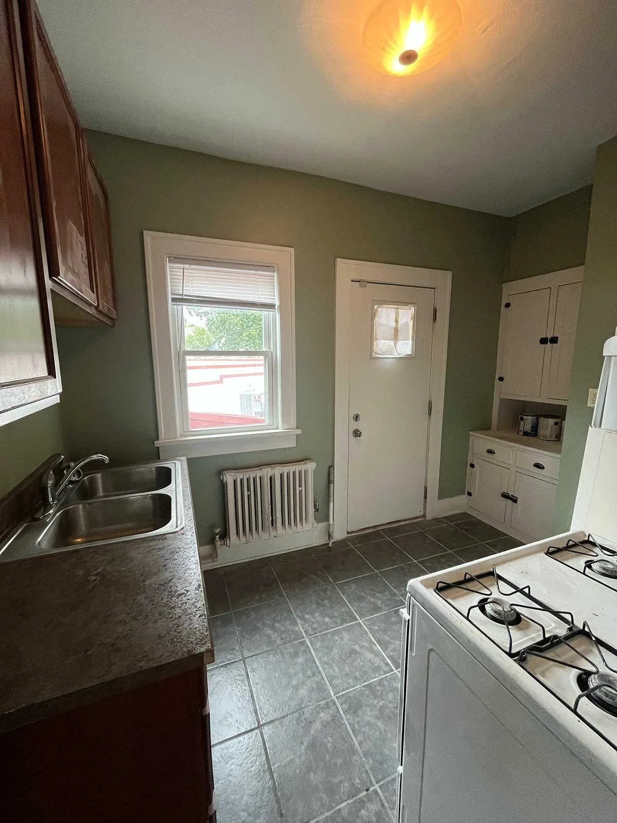 Small kitchen with green walls, a window with blinds, a white door, dark brown cabinets, a stove, a dual sink, and tiled flooring.