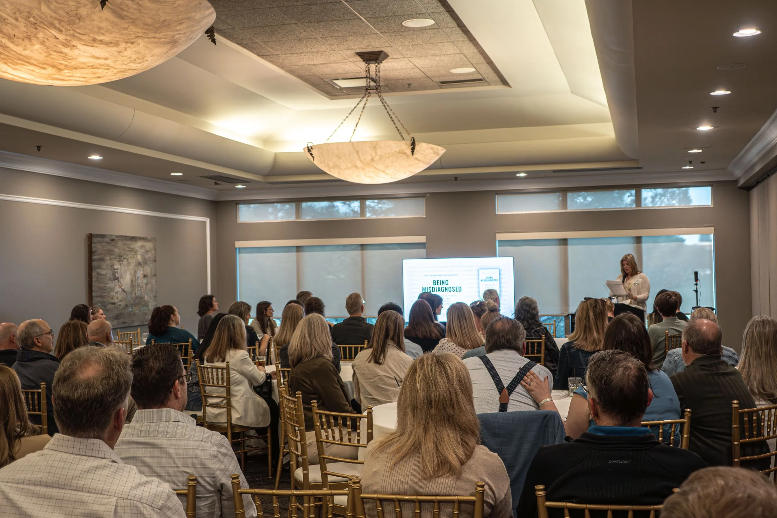 A conference room filled with seated attendees facing a woman at a podium giving a presentation. The room has large windows, ceiling lights, and decorative chandeliers.