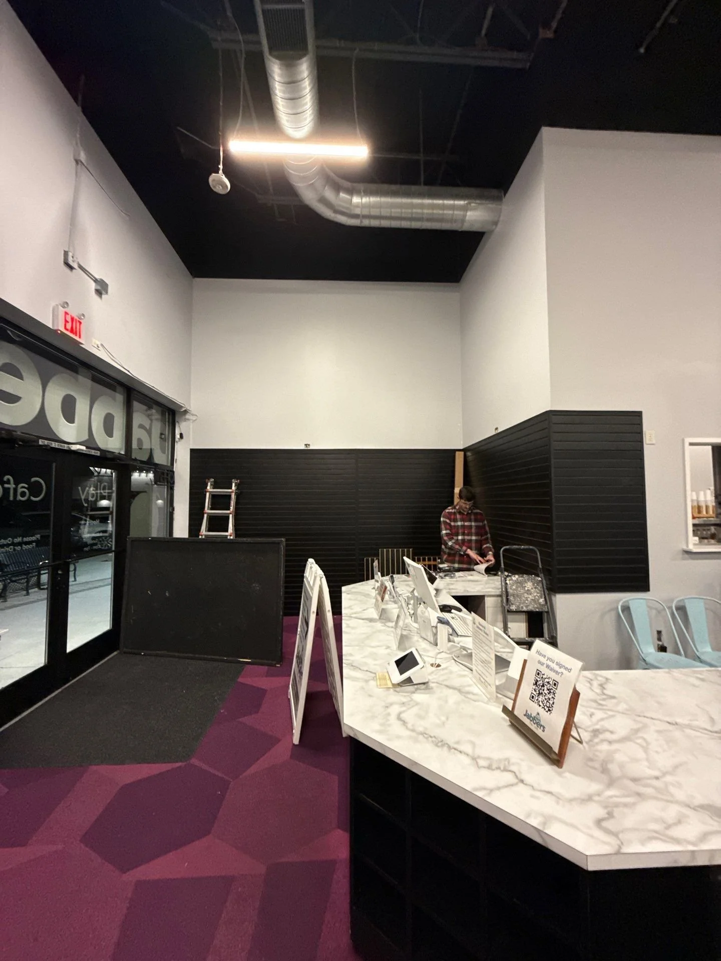 Interior of a modern office or lobby with a marble counter, a person in a red plaid shirt working at the desk, and a ladder leaning against the wall. The area has black and white walls, a high ceiling with exposed ductwork, and a large window or entrance on the left with