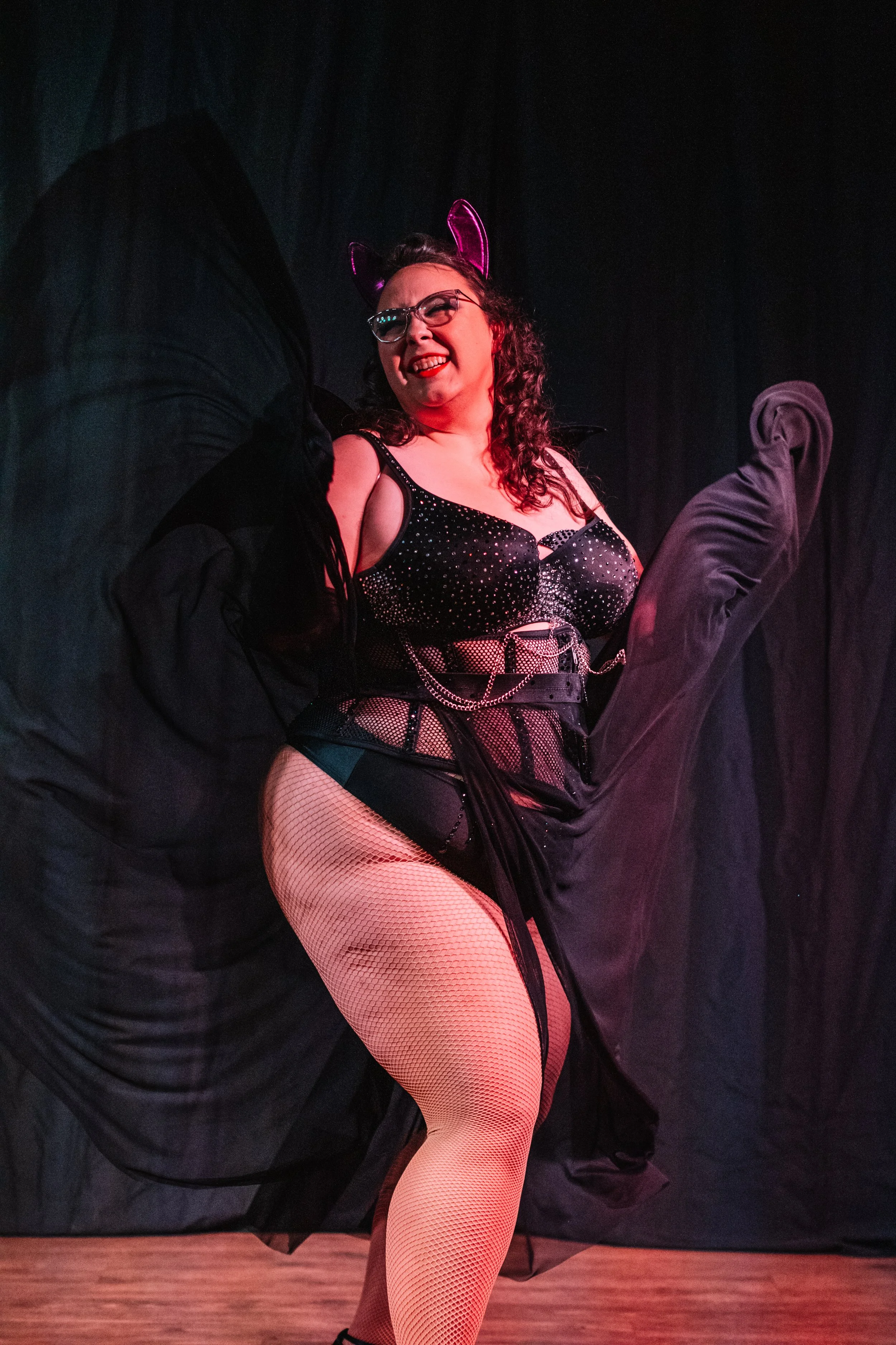 A woman performing a dance pose on stage, wearing a black sequined top, fishnet tights, and thick black pants, with cat ears headband and glasses, illuminated by stage lighting.
