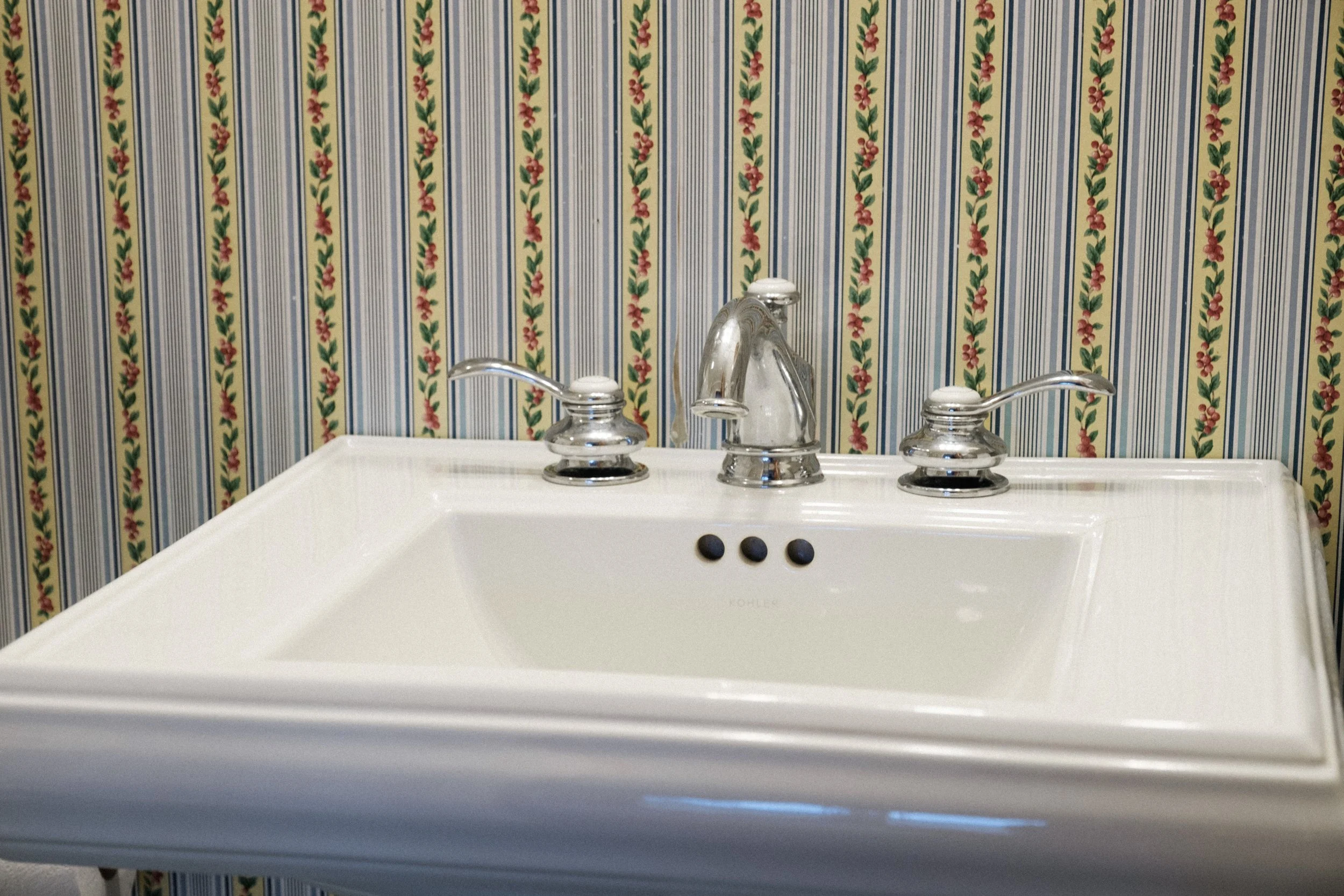 A white bathroom sink with a chrome faucet and two handles in front of a wallpaper with vertical stripes and floral patterns.
