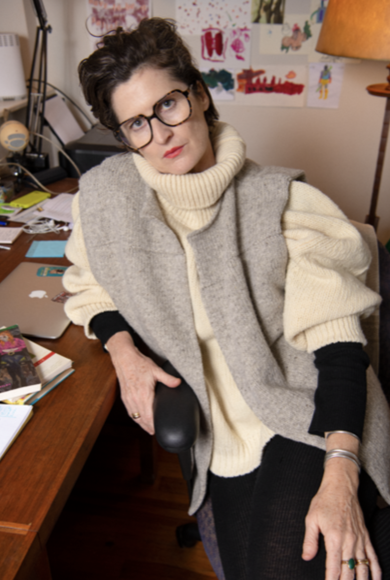 A woman with short brown hair, wearing glasses, a cream turtleneck sweater, and a gray sleeveless vest, sitting at a cluttered wooden desk in a home office with colorful children's artwork on the wall behind her and a table lamp to her right.