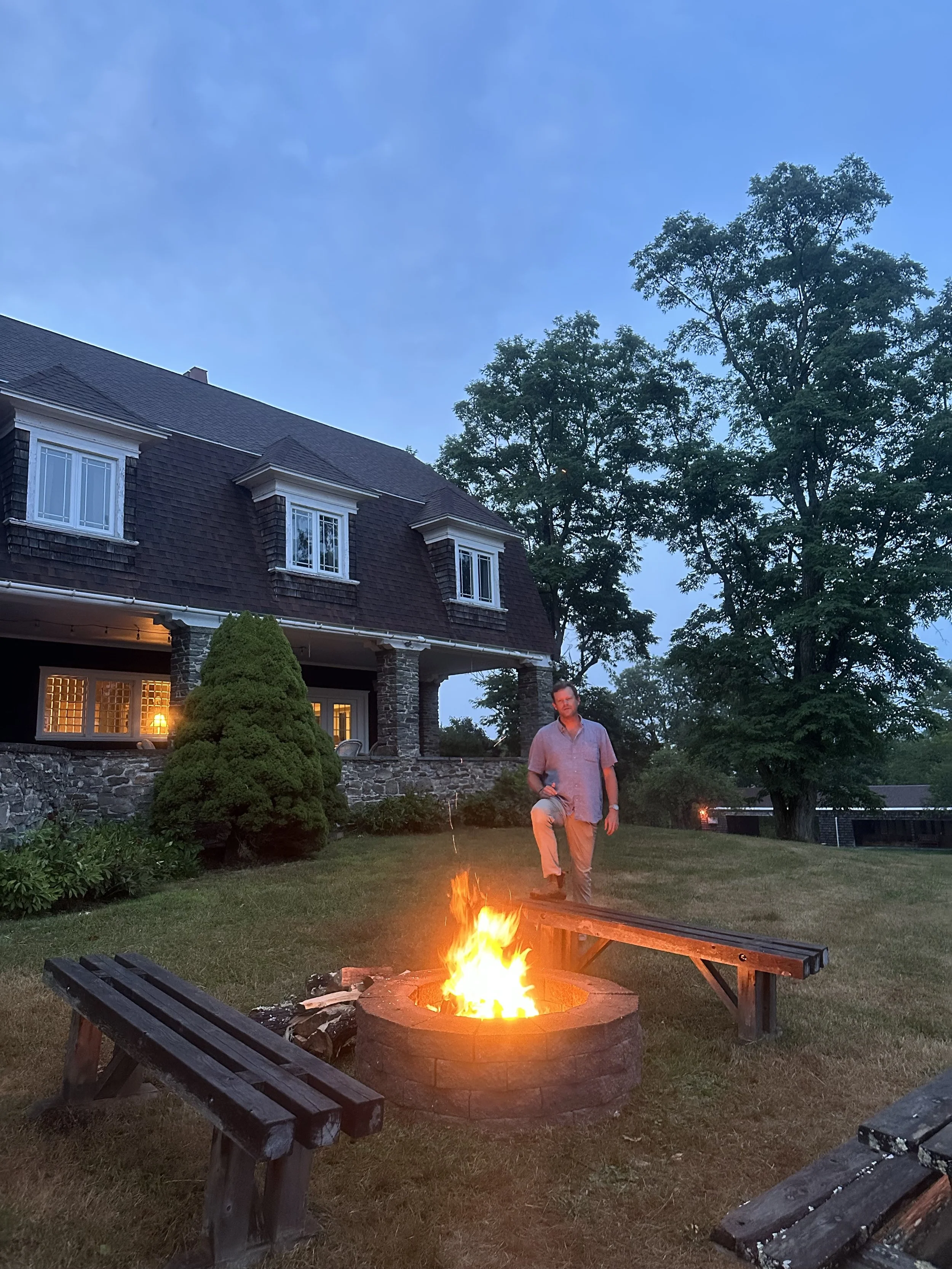 A man standing near a lit fire pit in a backyard during evening, with a large house in the background and trees surrounding the area.