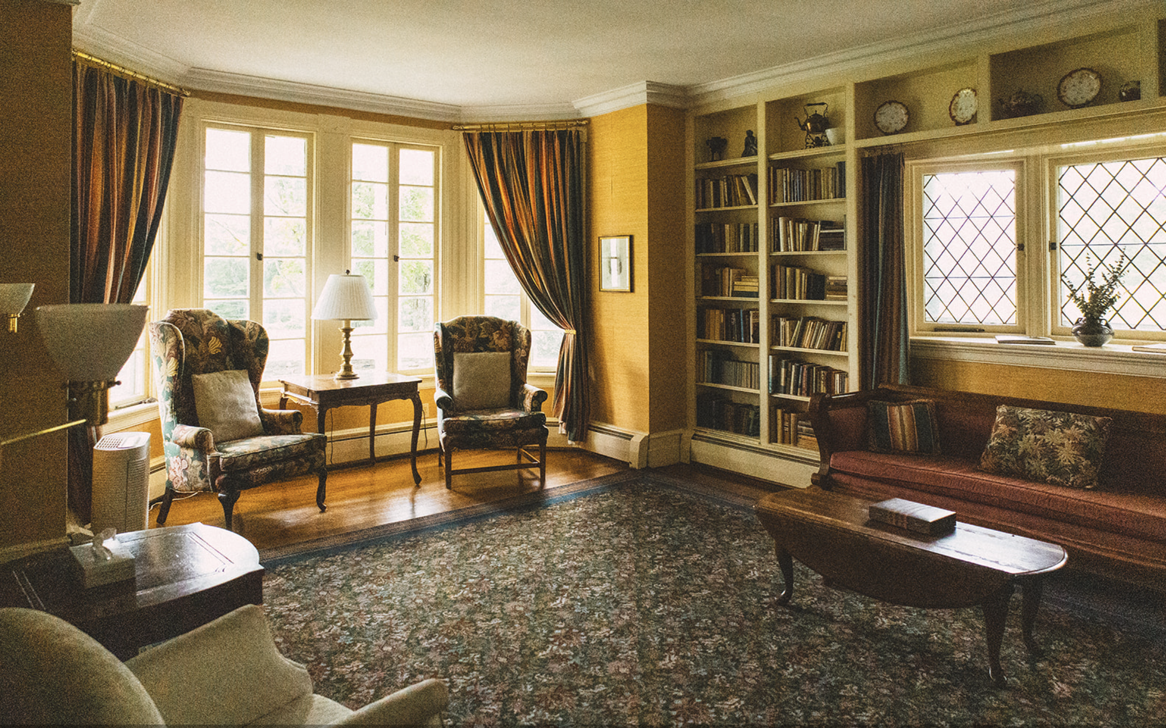 Living room with yellow walls, large windows with curtains, floral armchairs, a wooden table, a bookshelf, a sofa, a coffee table, and a patterned carpet.