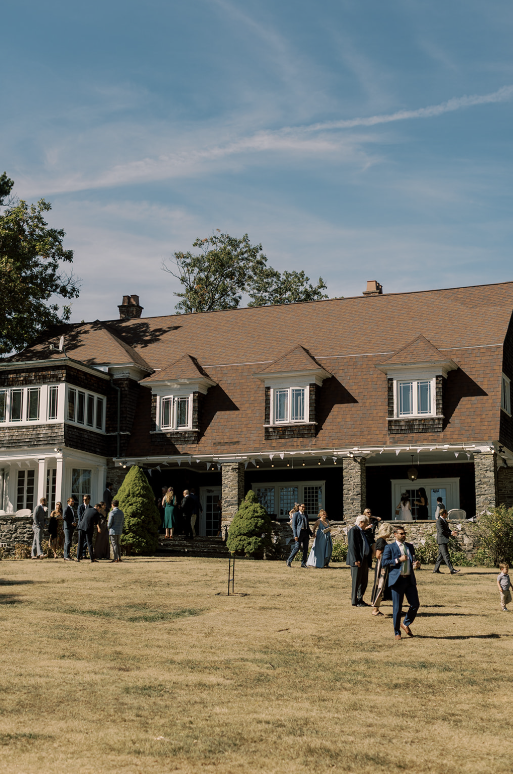 People gathered outside a large house with a brown shingle roof, stone porch columns, and multiple windows. The scene appears to be a social event on a sunny day.
