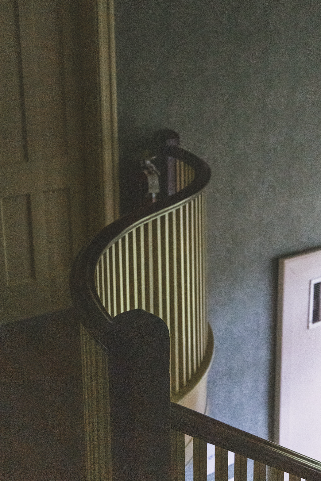 A person standing at the top of a wooden spiral staircase, partially obscured, wearing a black shirt and a face mask, holding a piece of paper or booklet.