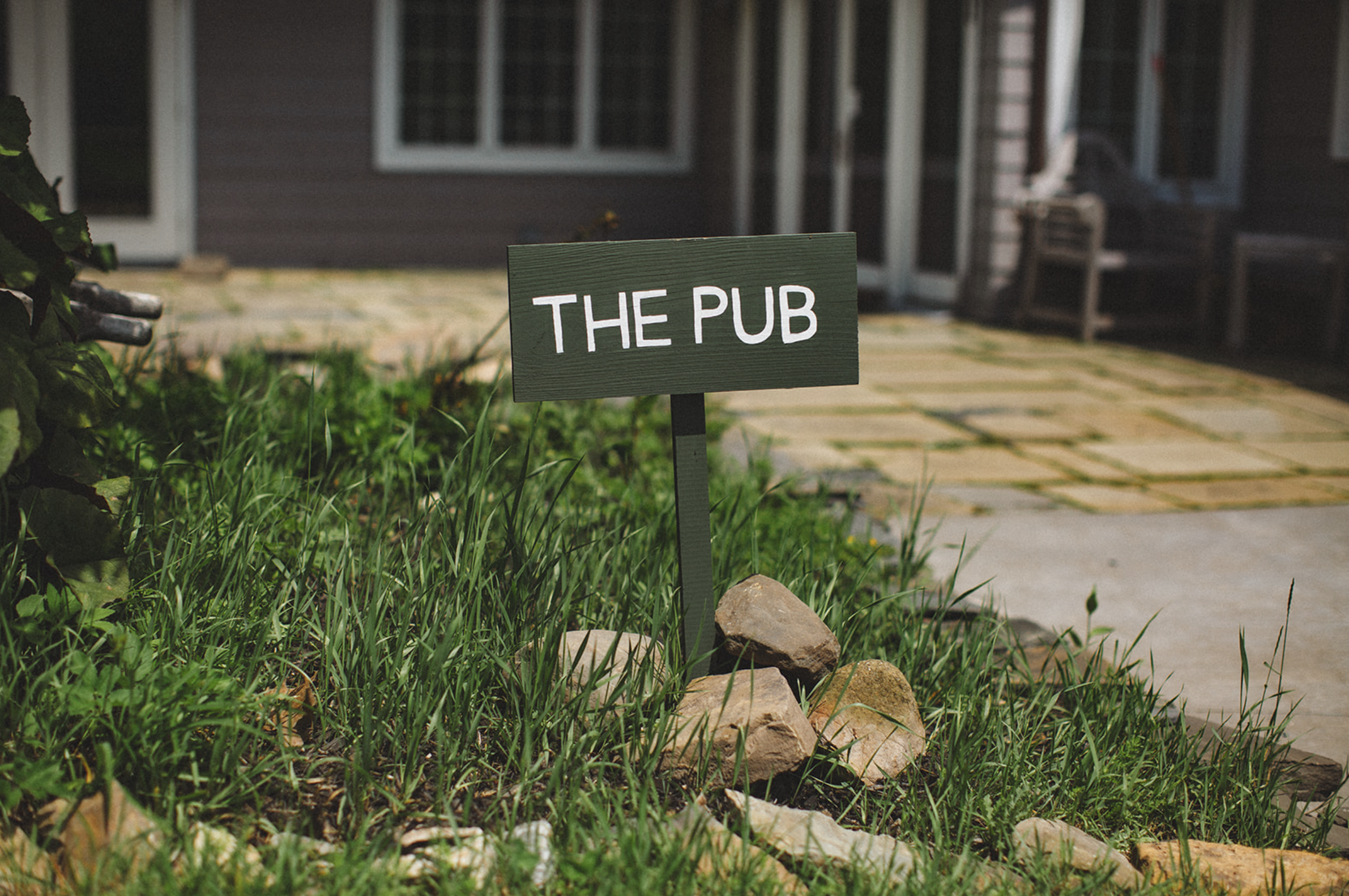 A green wooden sign reading 'THE PUB' is placed among rocks in a grassy garden area outside a house with patio stones and a barbecue, with the house's dark exterior and windows in the background.