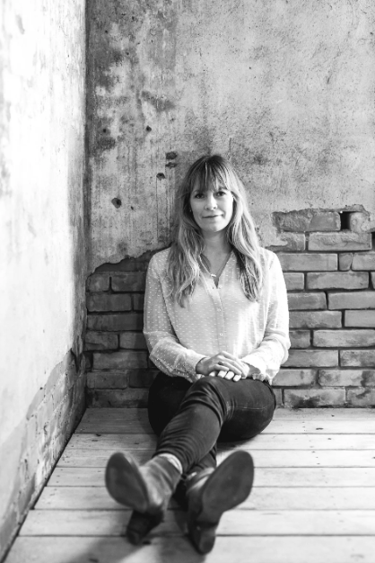 A young woman sitting on a wooden floor in front of a rough concrete and brick wall, looking at the camera.