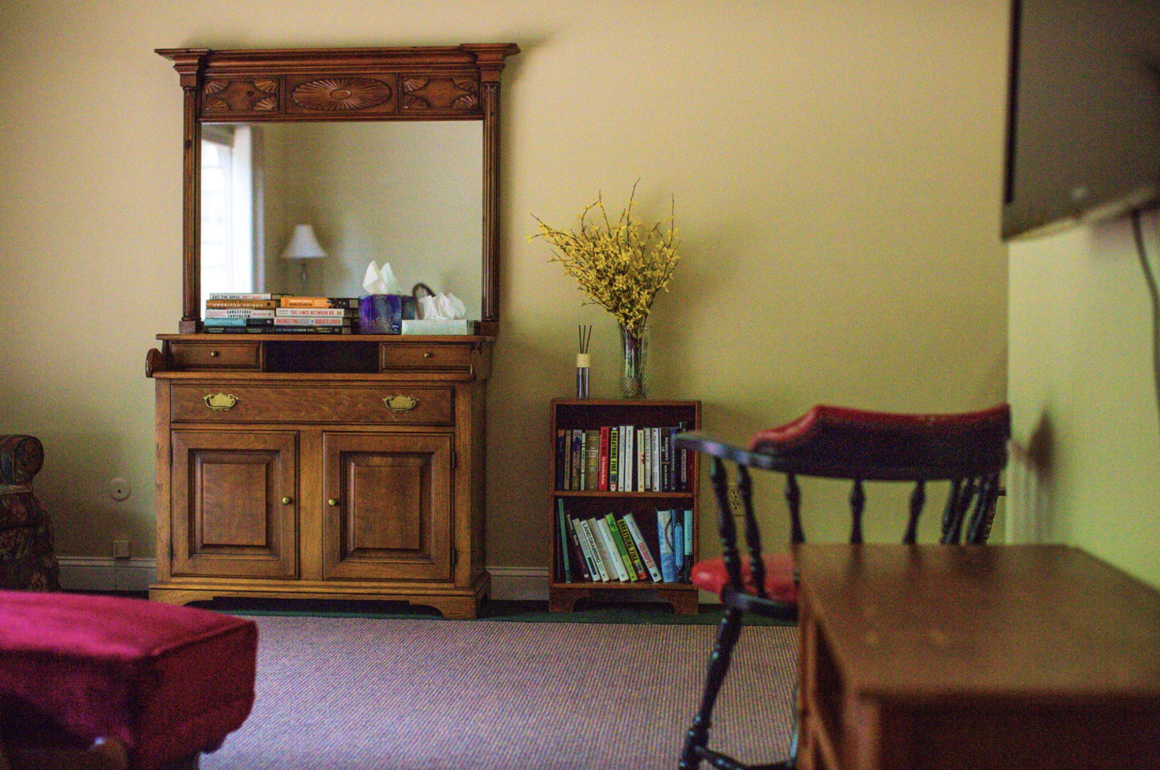 Living room corner with a wooden dresser and mirror, a small bookshelf with books and yellow flowers, and a chair with a red cushion.