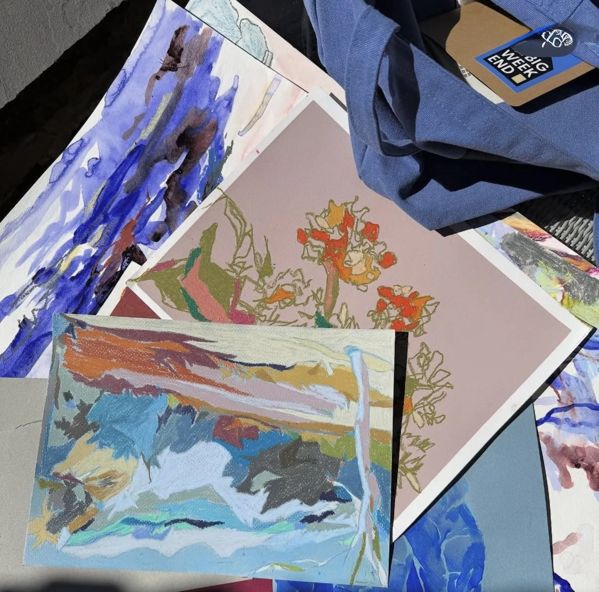 Several colorful pieces of abstract artwork and drawings are spread out on a surface, with a blue backpack and a conference badge nearby.