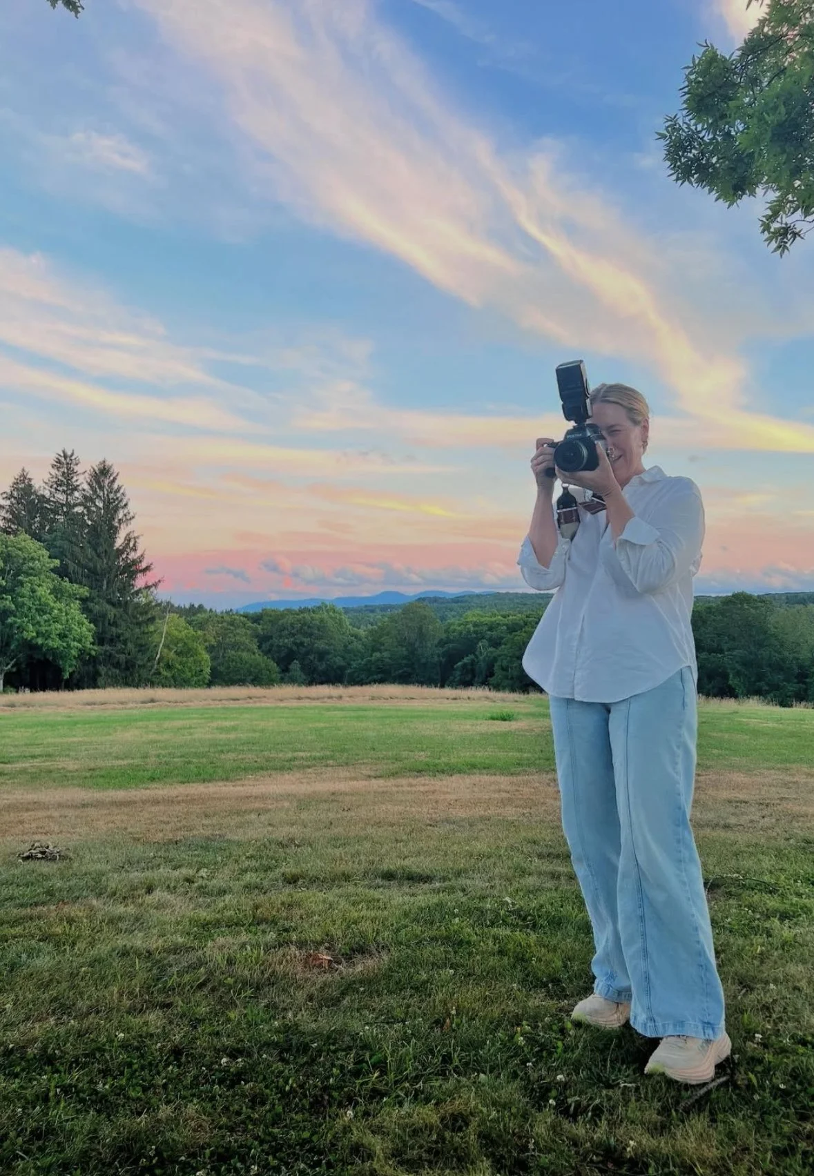 A woman in white shirt and light blue jeans standing on a grassy field, taking a photograph with a camera, with trees and a colorful sunset sky in the background.