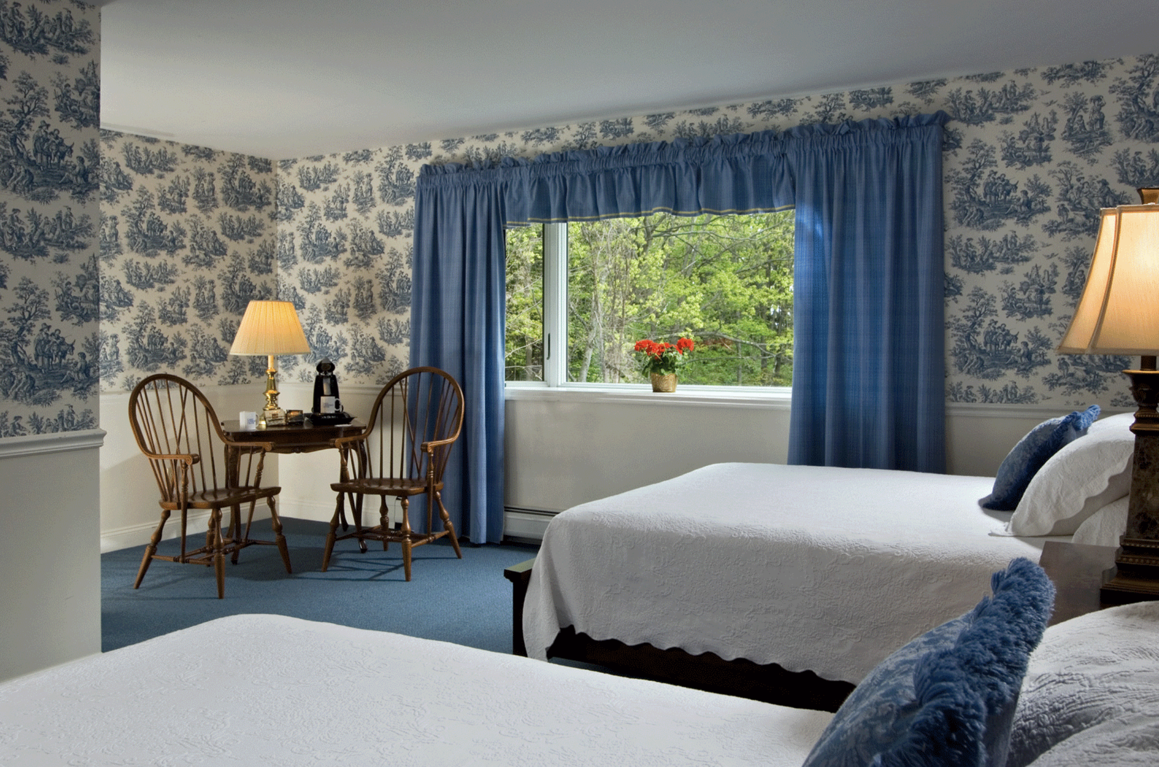 A cozy hotel room with two beds dressed in white quilts, blue pillows, and matching curtains. There is a window with lush green trees outside and a small flower pot on the windowsill. A small wooden table with two chairs, a lamp, and a coffee maker c