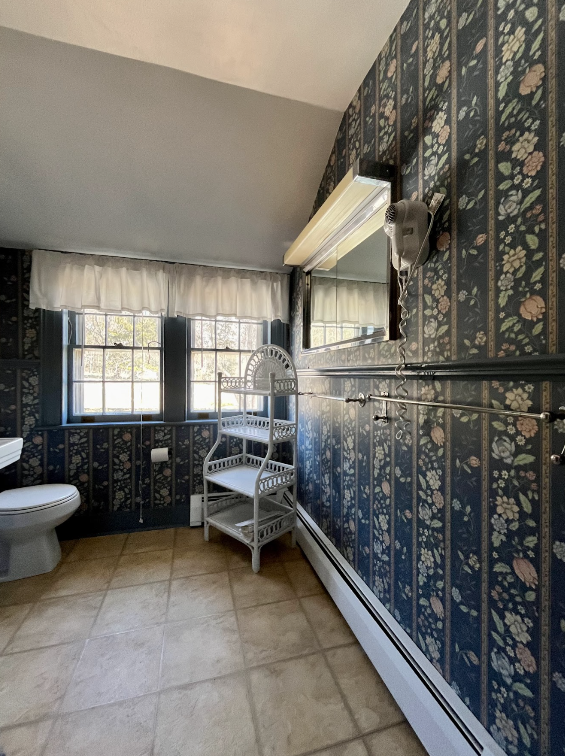 A bathroom with beige tiled floor, floral wallpaper, a window with white curtains, a white toilet, a white wicker shelf, and a mirror on the wall.