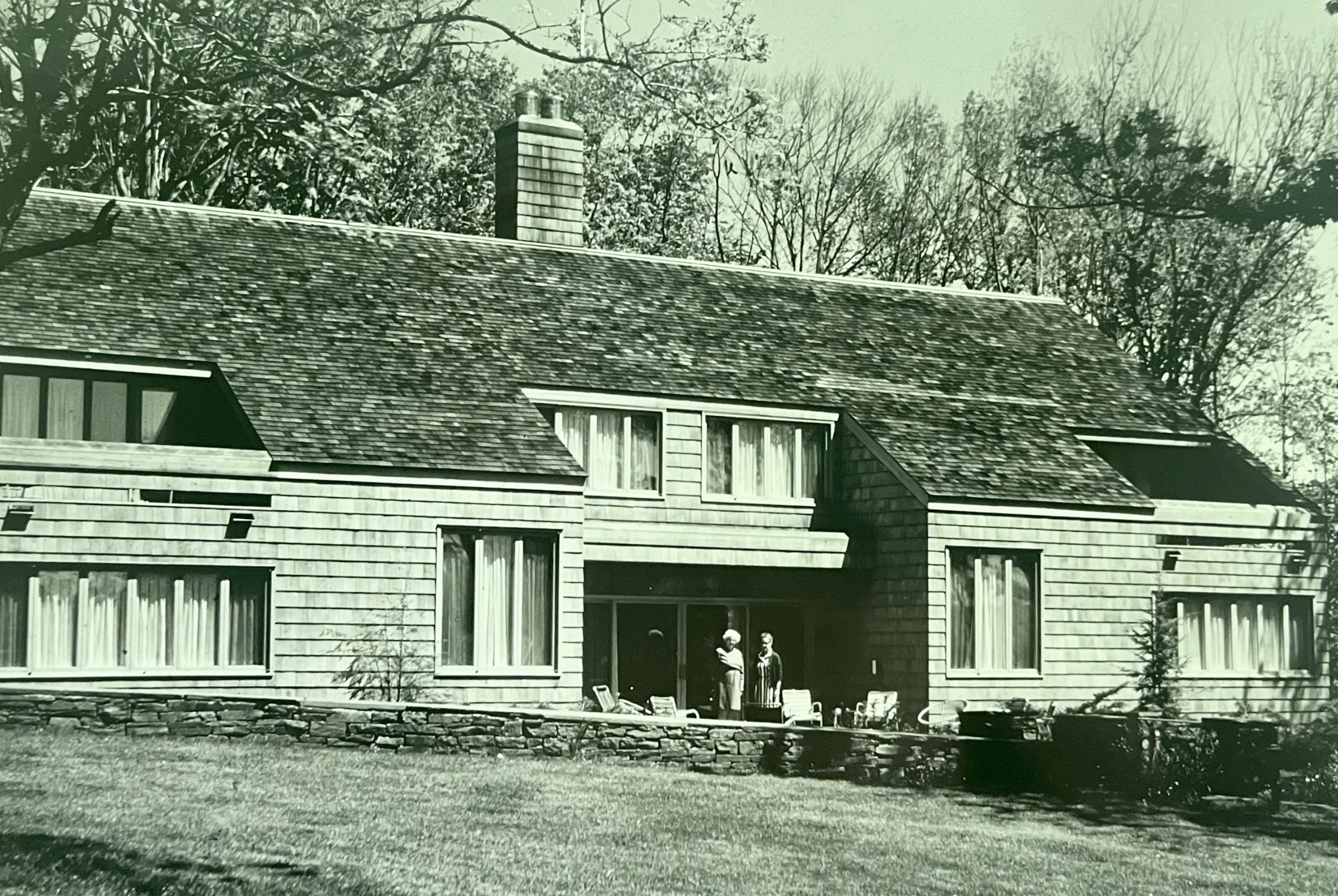 A large house with a sloped roof, multiple windows with curtains, and a stone foundation. Two women are standing on the porch, and there are several chairs in front of the house. The photo appears to be old, with a green tint and trees in the backgro