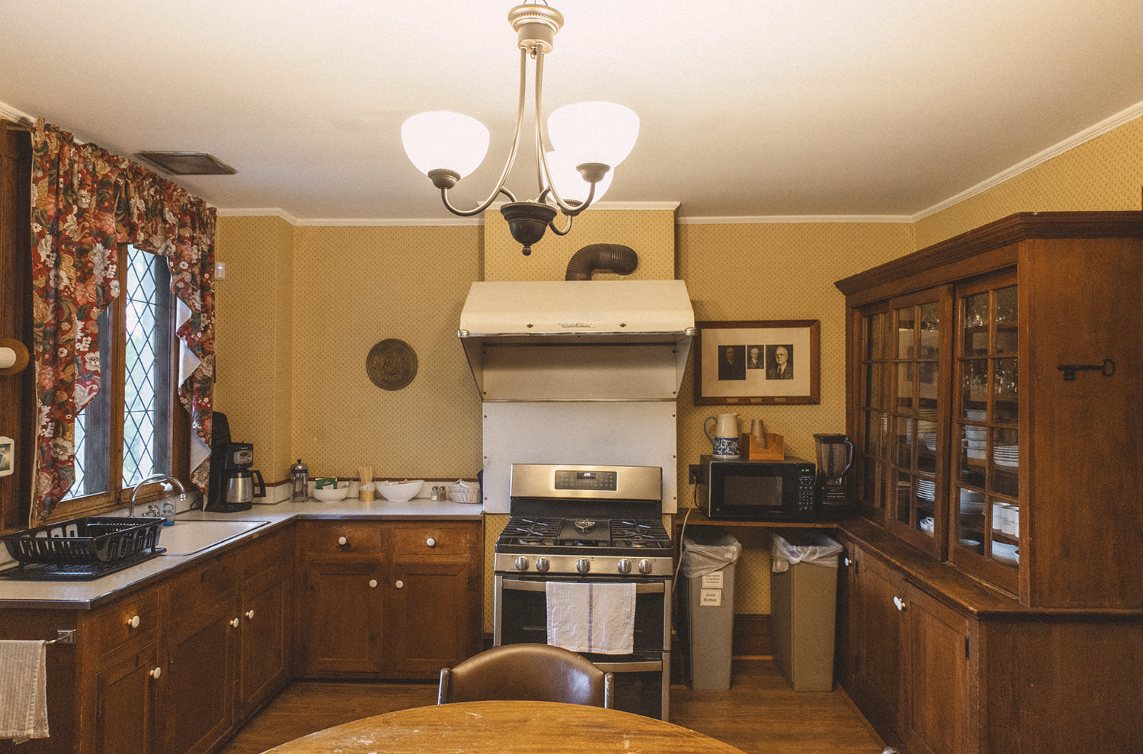 Traditional kitchen with wooden cabinets, yellow polka dot wallpaper, a window with floral curtains, a stove with a vent hood, a dining table in the foreground, and various appliances including a microwave, coffee maker, and a blender.