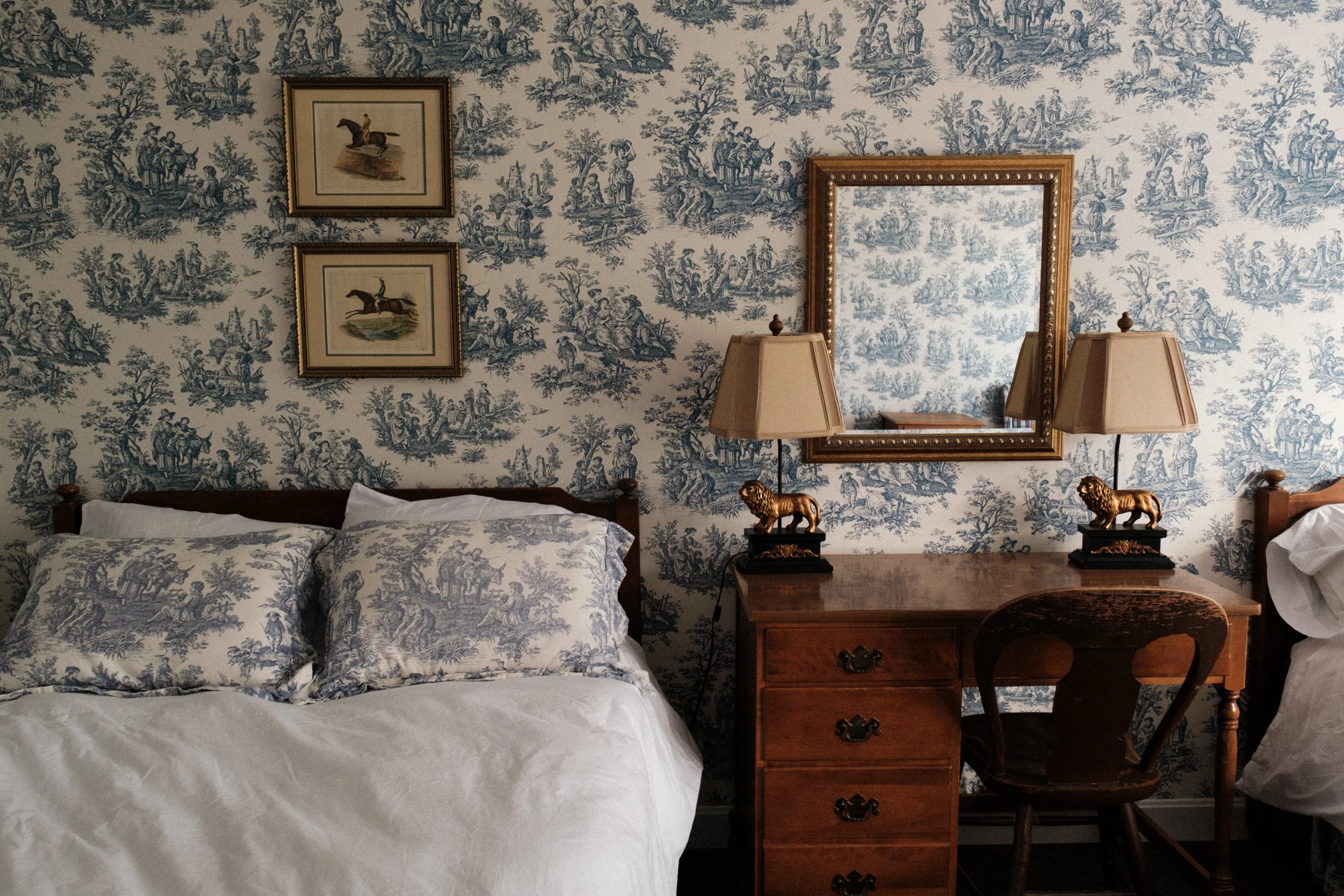 A bedroom with blue and white toile wallpaper, the bed has pillows matching the wallpaper, a wooden desk with two decorative lion lamps, a mirror above the desk, and framed horse pictures on the wall.