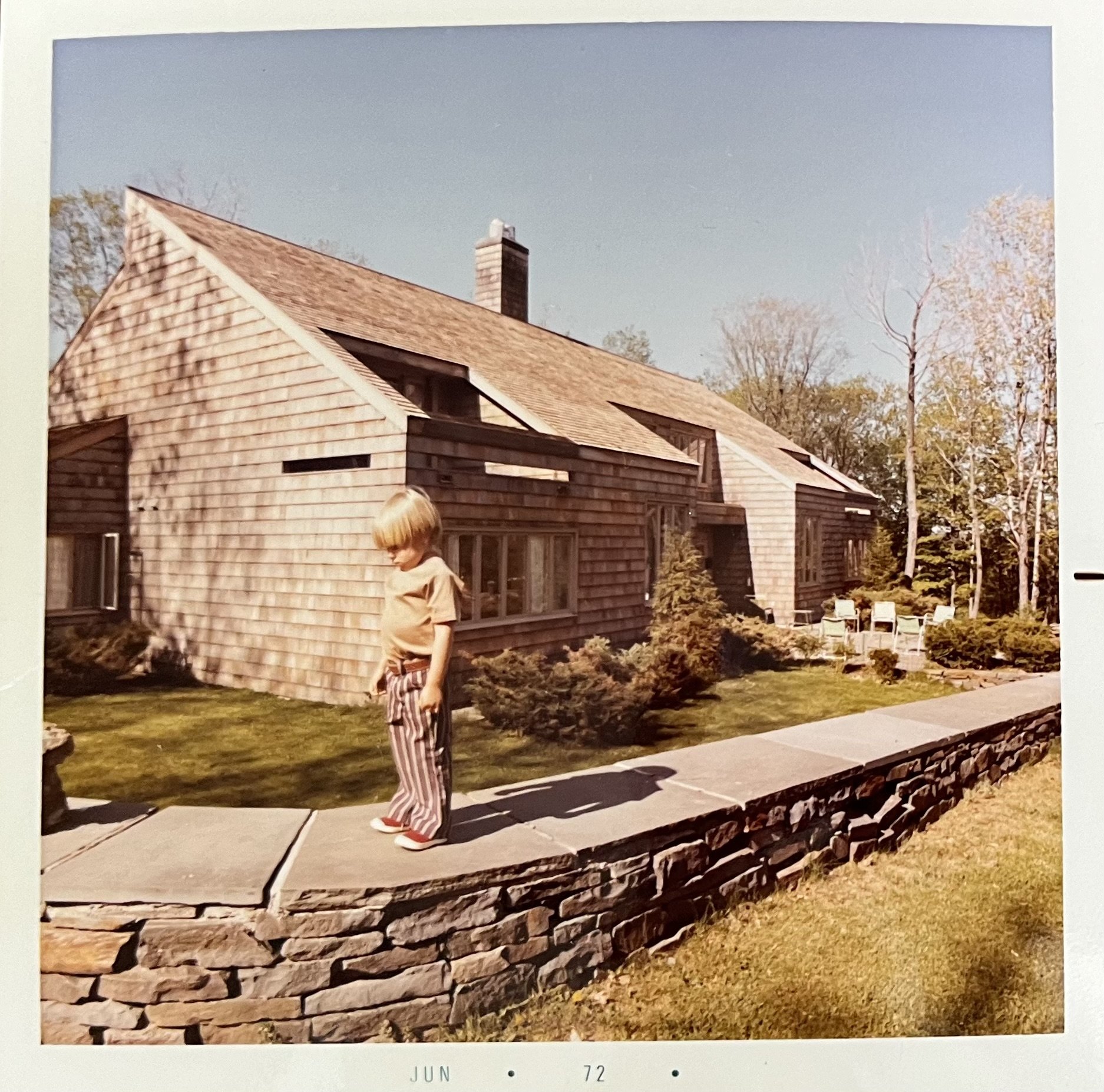 A young boy with blonde hair wearing a beige shirt and striped pants stands on a stone wall in front of a wood-shingled house with large windows, surrounded by trees and a manicured lawn.