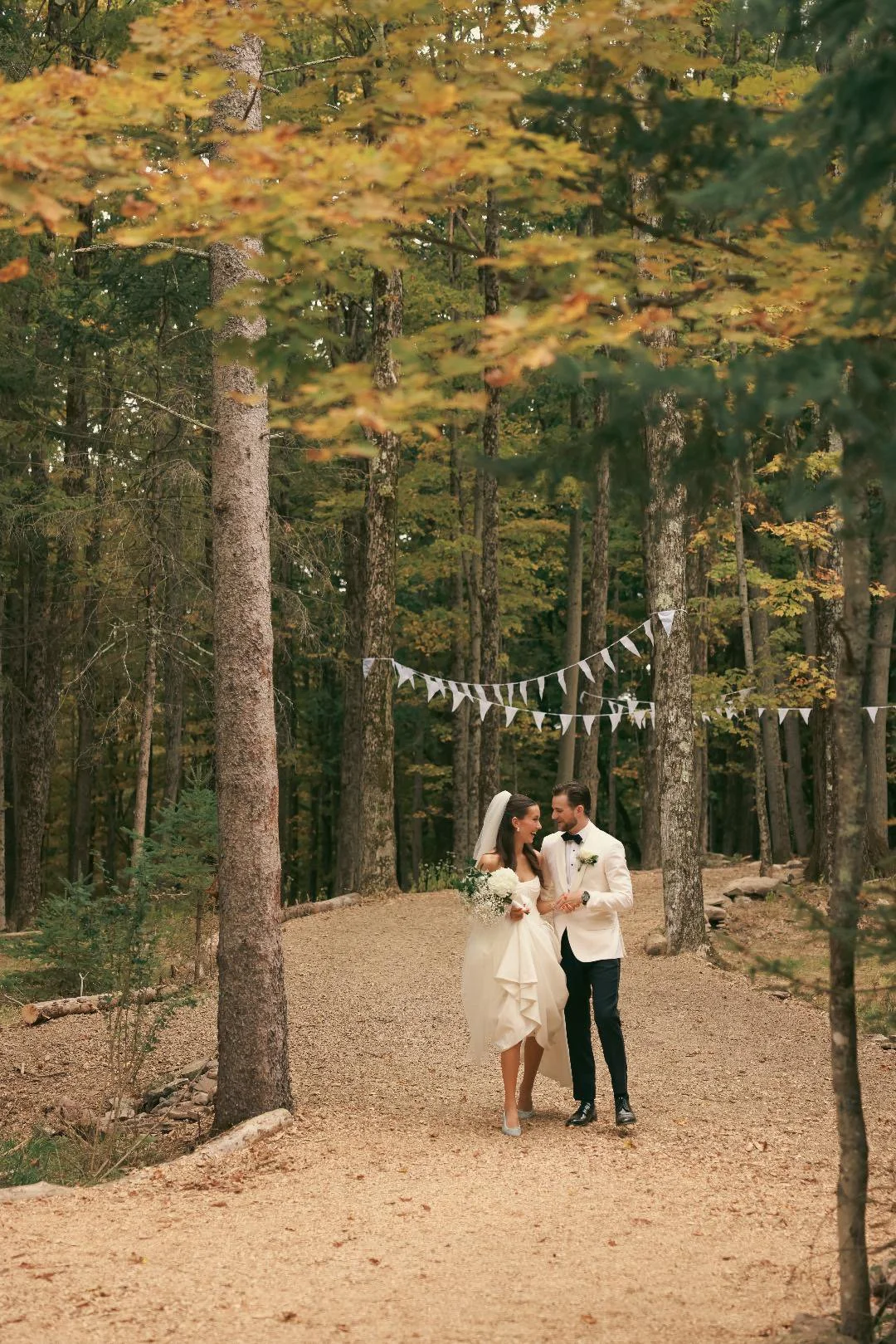 Bride and groom walking together in a forest with autumn foliage, holding hands and smiling, with white bunting hanging between trees above them.
