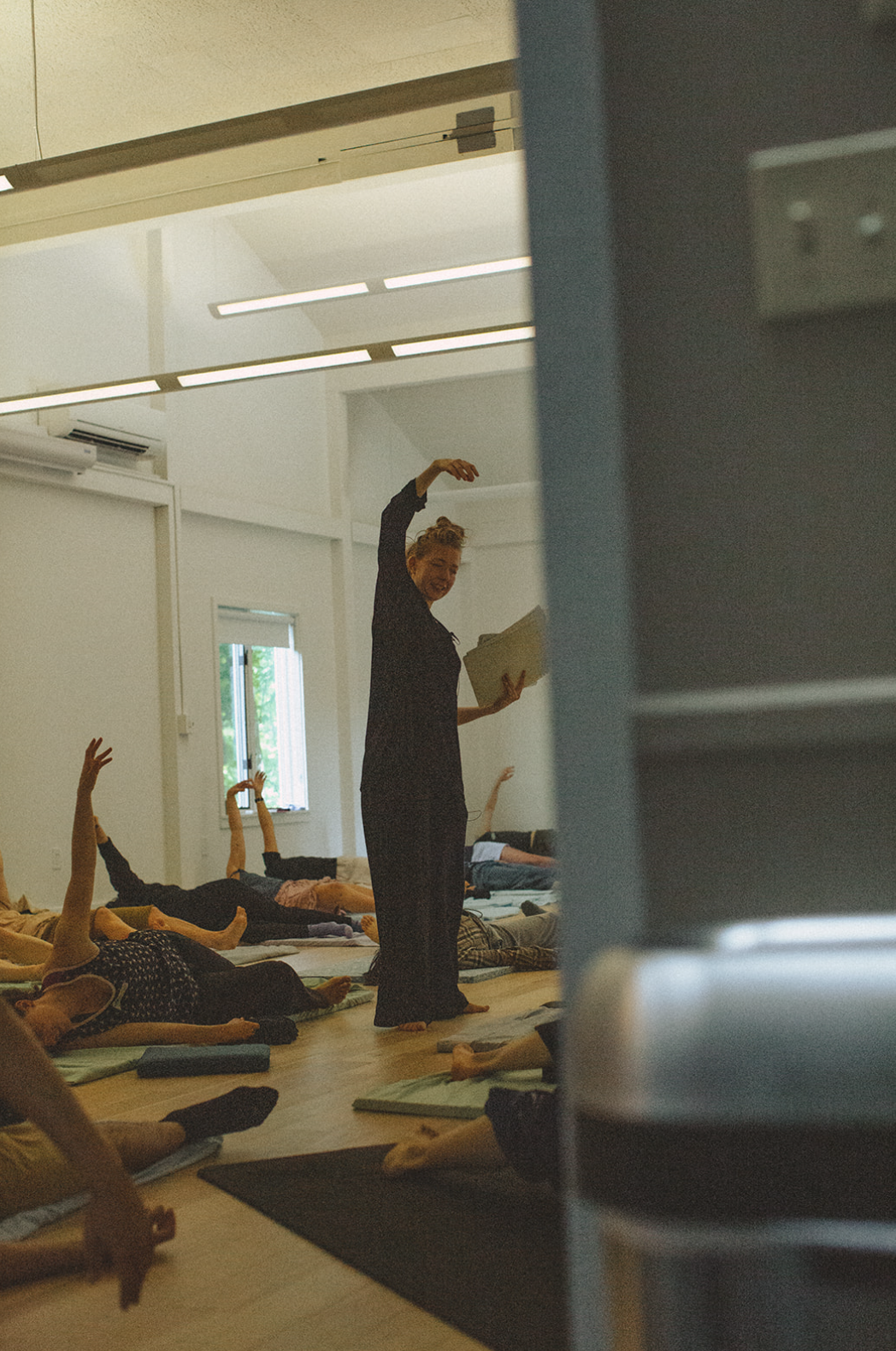 People participating in a yoga or meditation class, lying on mats with some raising their hands, led by an instructor holding a book and smiling.