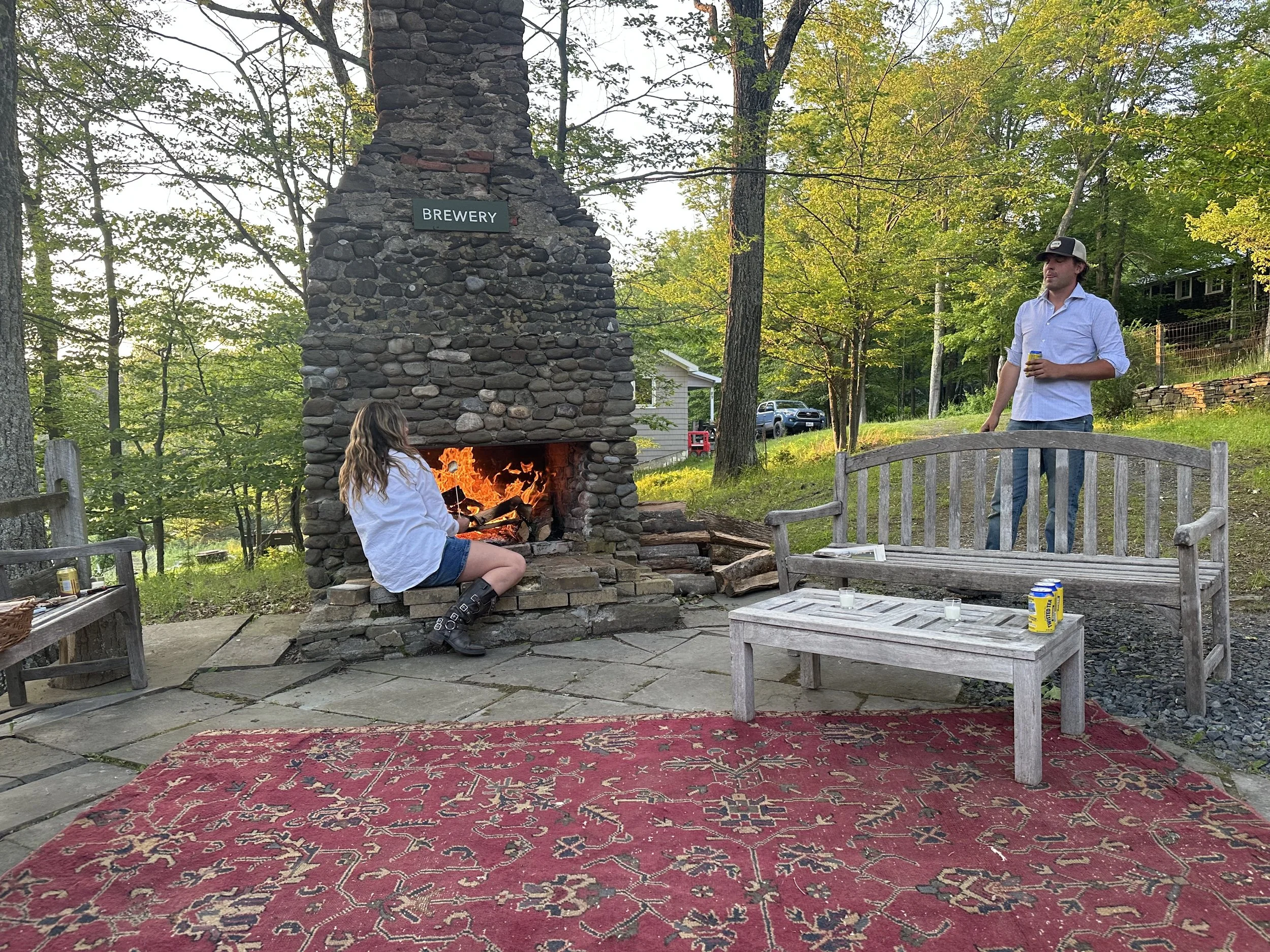 A girl seated on a stone ledge in front of a stone fireplace with a fire burning inside, outdoors in a wooded area. A man standing nearby holds a drink. There are drinks and candles on a wooden bench and table. A red patterned rug covers part of the 