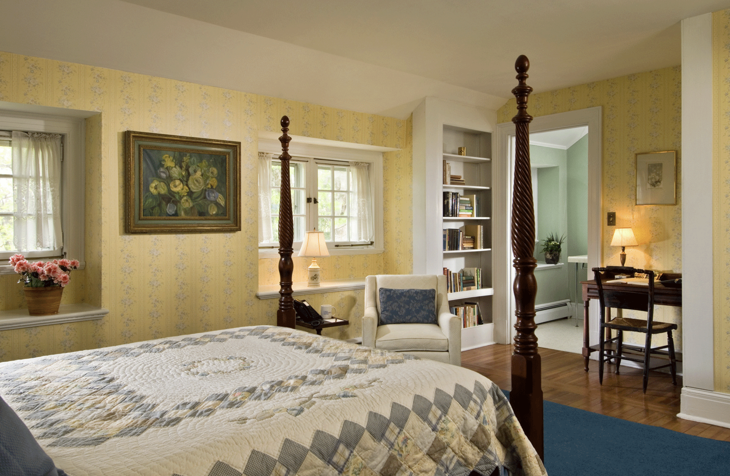 A cozy bedroom with yellow floral wallpaper, a four-poster bed with quilts, a window with white curtains, potted flowers, an armchair with a blue pillow, a bookshelf filled with books, a desk with a lamp, and a doorway leading to a green-colored room