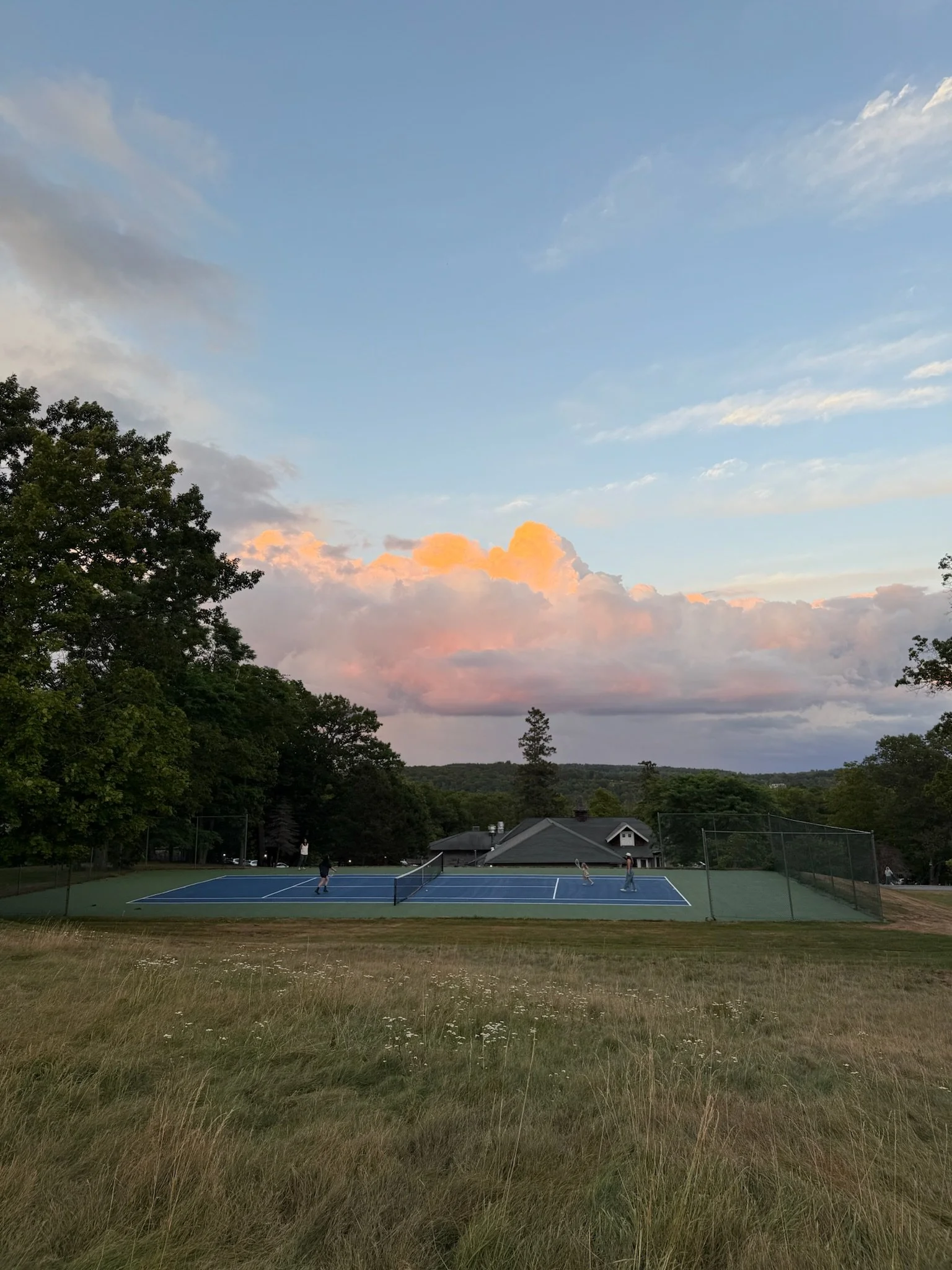 A grassy field with a blue tennis court surrounded by trees. Three people are playing tennis on the court. The background shows a house and a cloudy sky with pink and orange clouds during sunset.