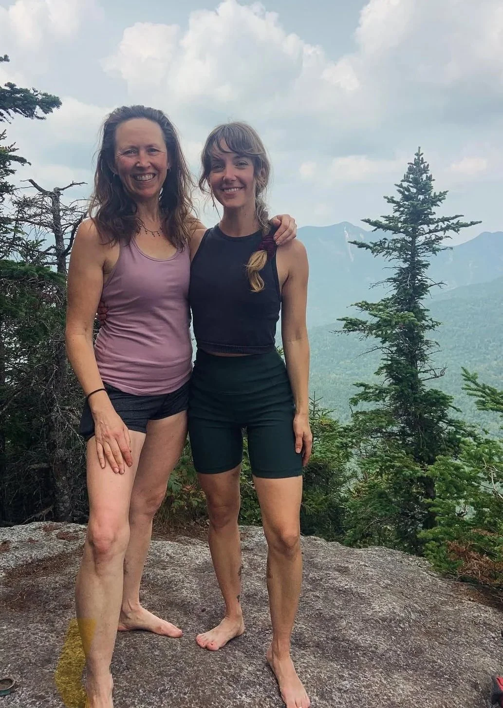 Two women standing on a rock with trees and mountain scenery behind them, smiling, wearing athletic clothing, with one woman in a purple tank top and black shorts, and the other in a black tank top and black shorts.