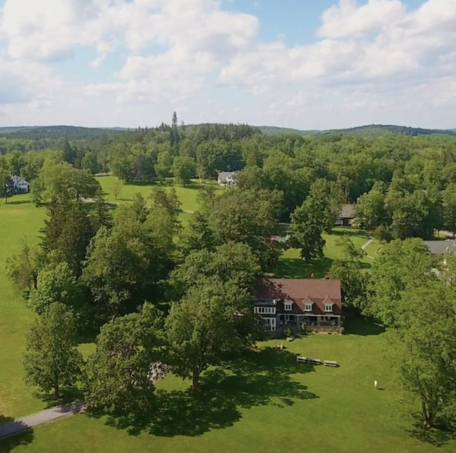 Aerial view of a green landscape with trees, houses, and rolling hills under a partly cloudy sky.