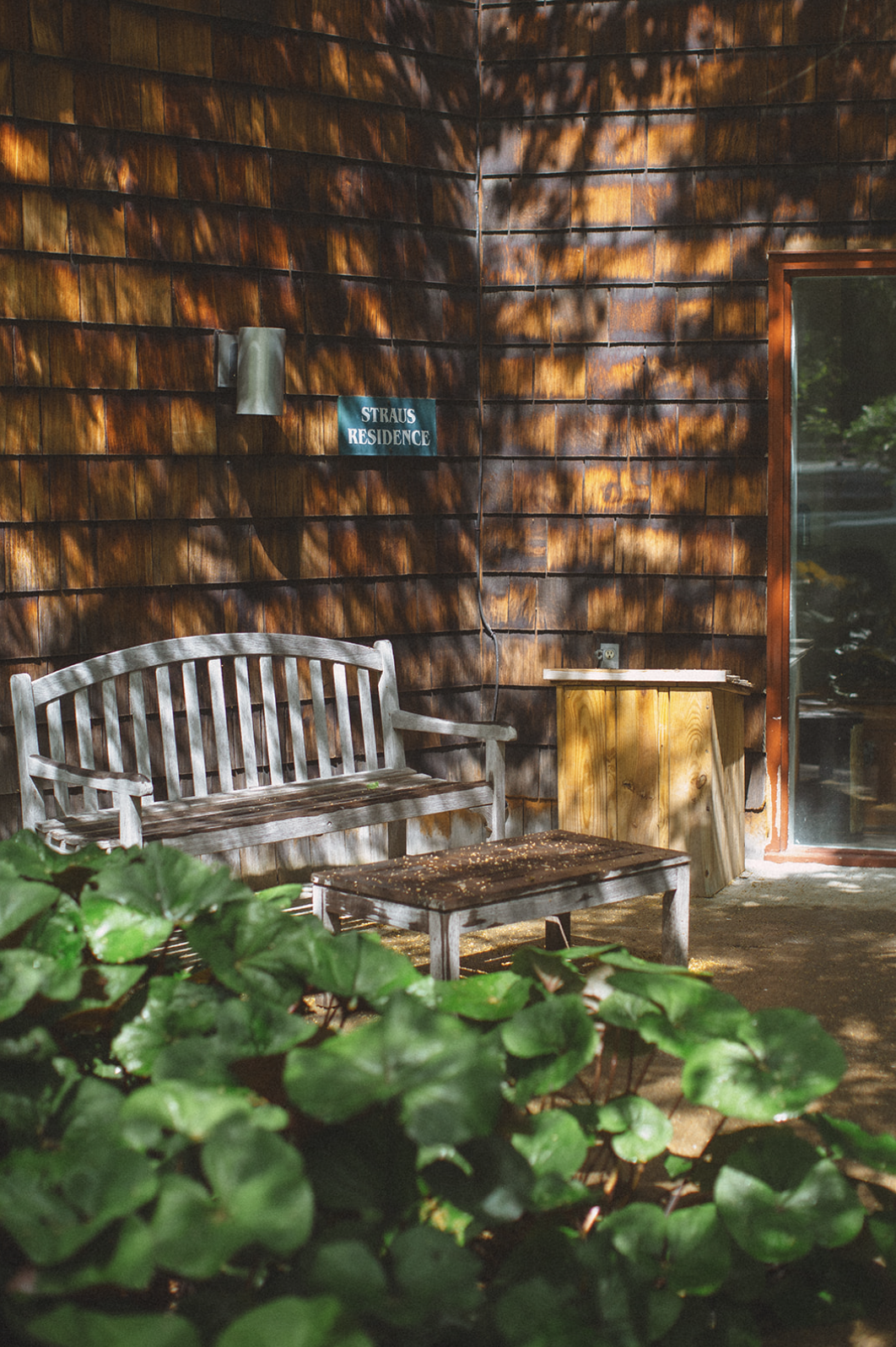 A wooden bench and table outside a house with shingle siding, a small wooden cabinet, and a sign reading 'STRAUS RESIDENCE' on the wall surrounded by greenery and dappled sunlight.