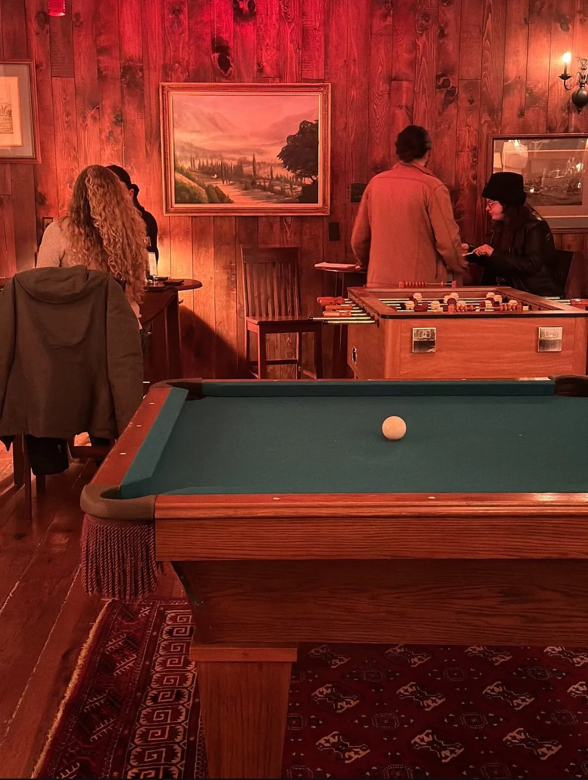 A bar or game room with a pool table in the foreground and a foosball table in the background. There are several people around, some standing and some sitting. The room has wooden walls decorated with paintings and dim lighting, with a small wall sconce visible on the right.