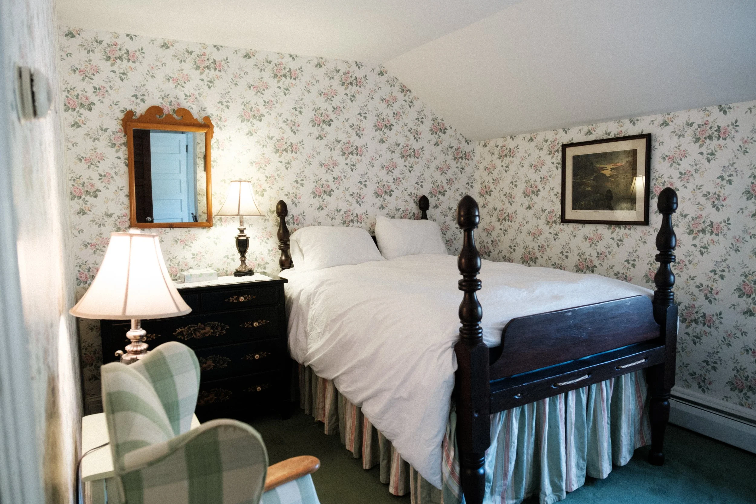 A cozy bedroom with a large four-poster bed, floral wallpaper, a mirror, lamps, and a framed painting on the wall.