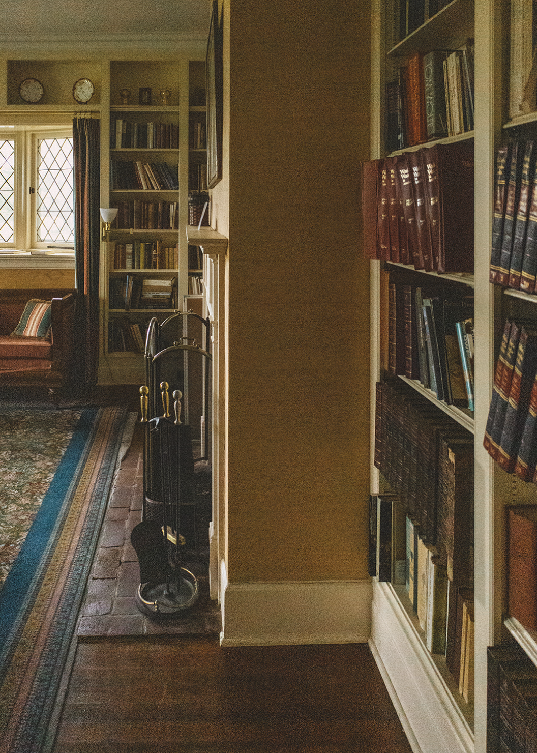 A cozy living room with large windows, a bookshelf with books, a fireplace with fireplace tools, a colorful patterned rug, and a sofa with striped cushions.